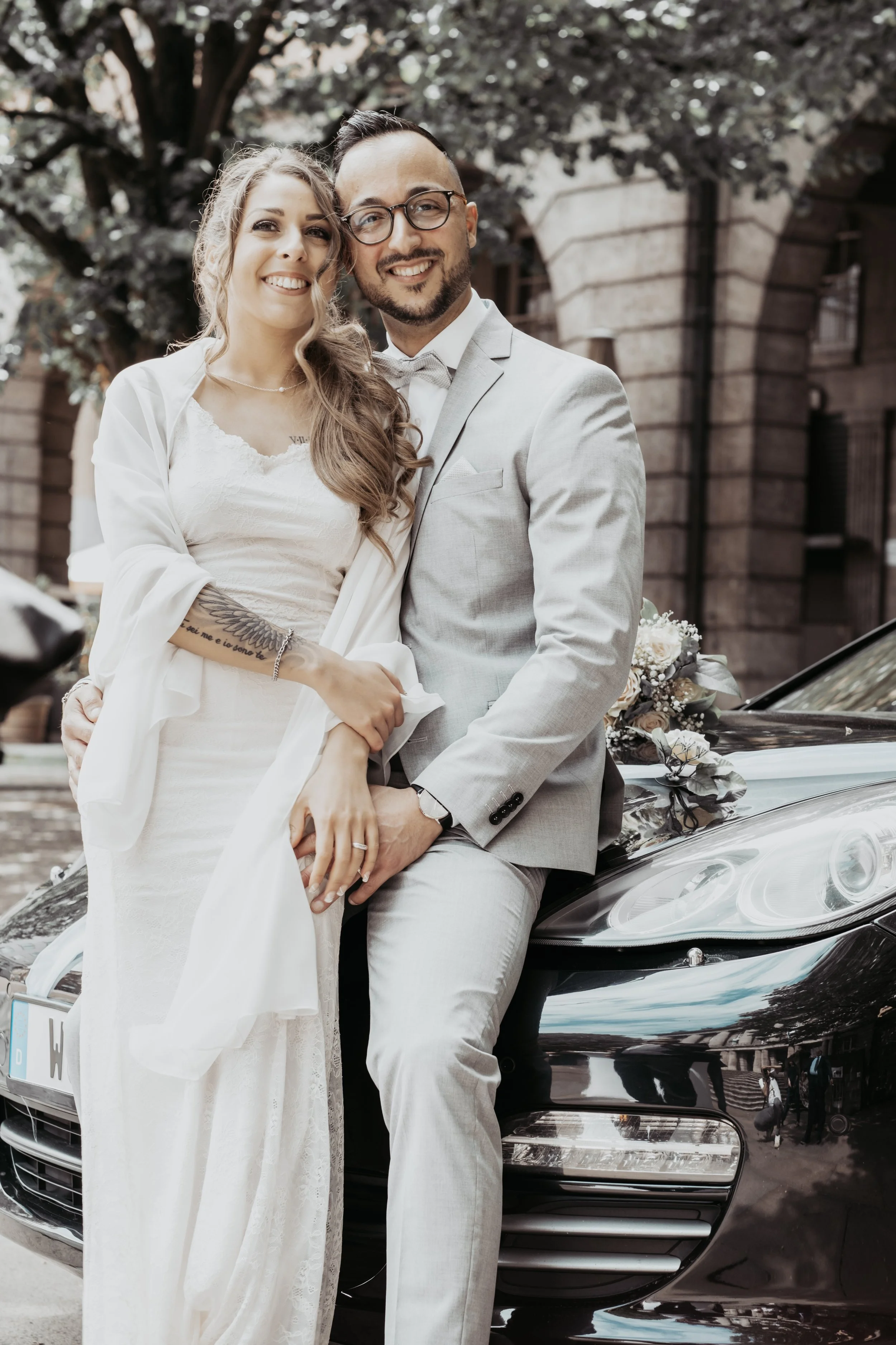 A bride and groom sitting on the hood of a black car, smiling and holding hands, outdoors with trees and historic buildings in the background.