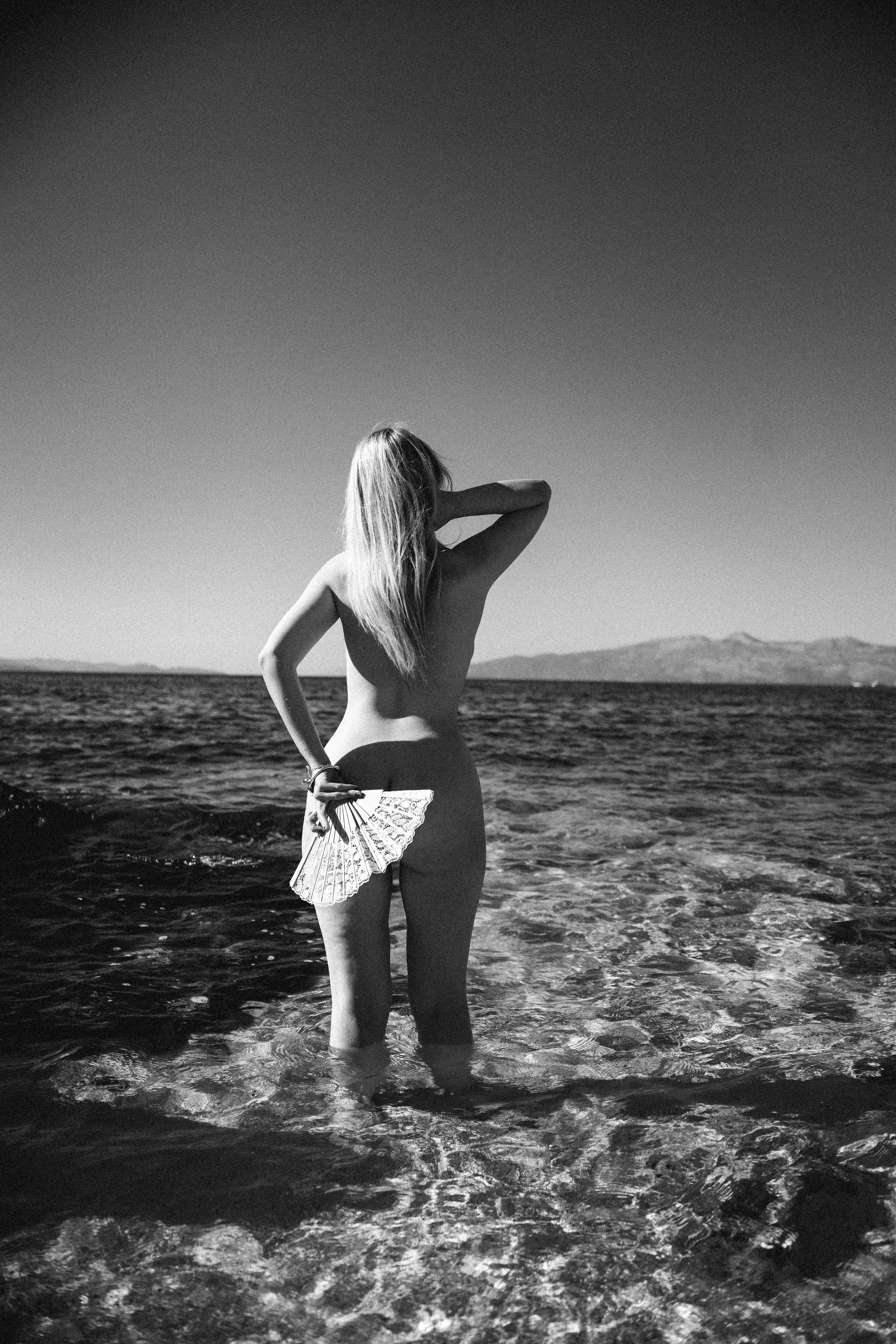 A woman with long hair standing in the water at a beach, holding a decorative fan in her hand, with mountains in the background and a clear sky.