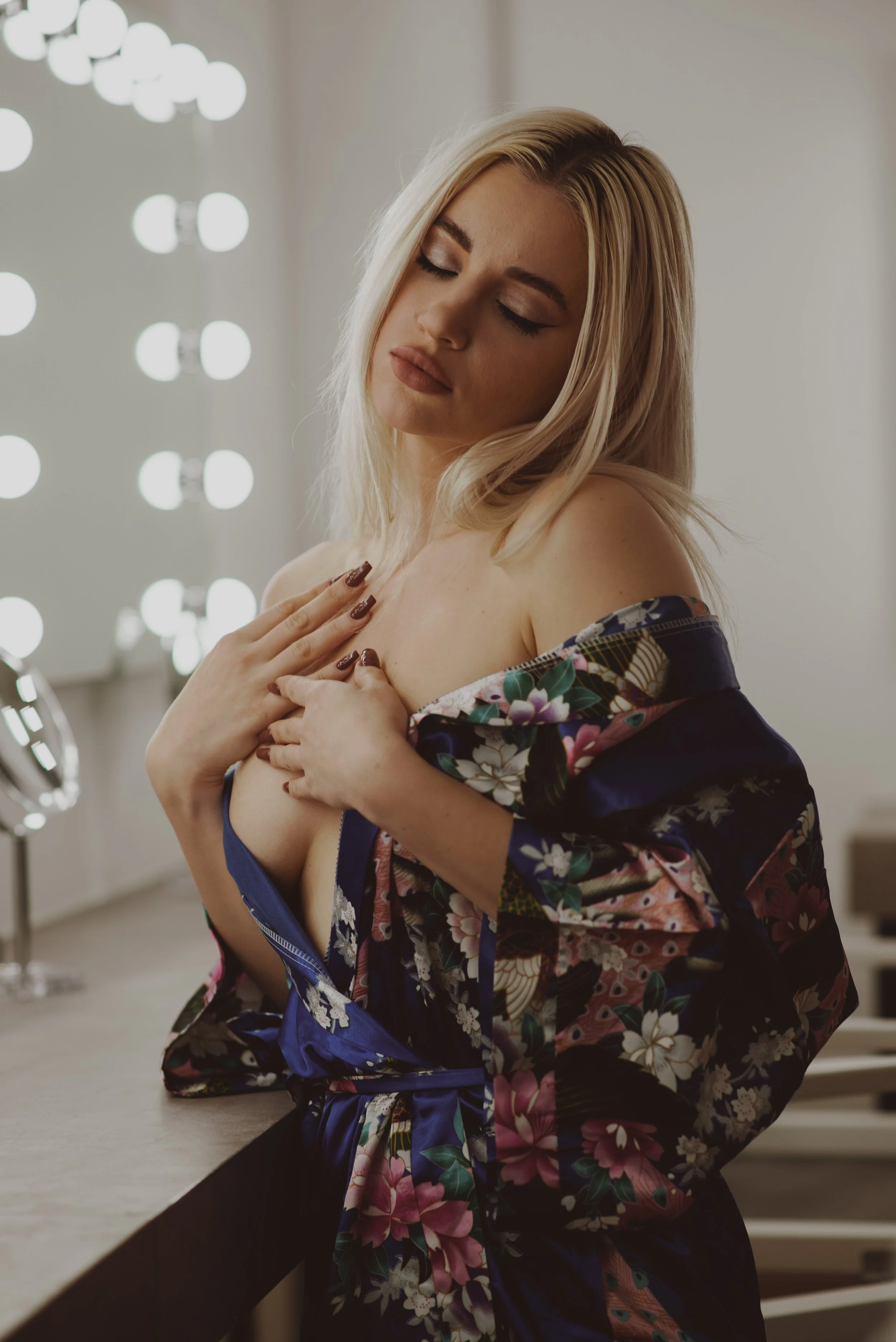A woman with blonde hair and makeup, wearing a floral kimono, touching her chest with her eyes closed, in a dressing room with bright lights in the background.