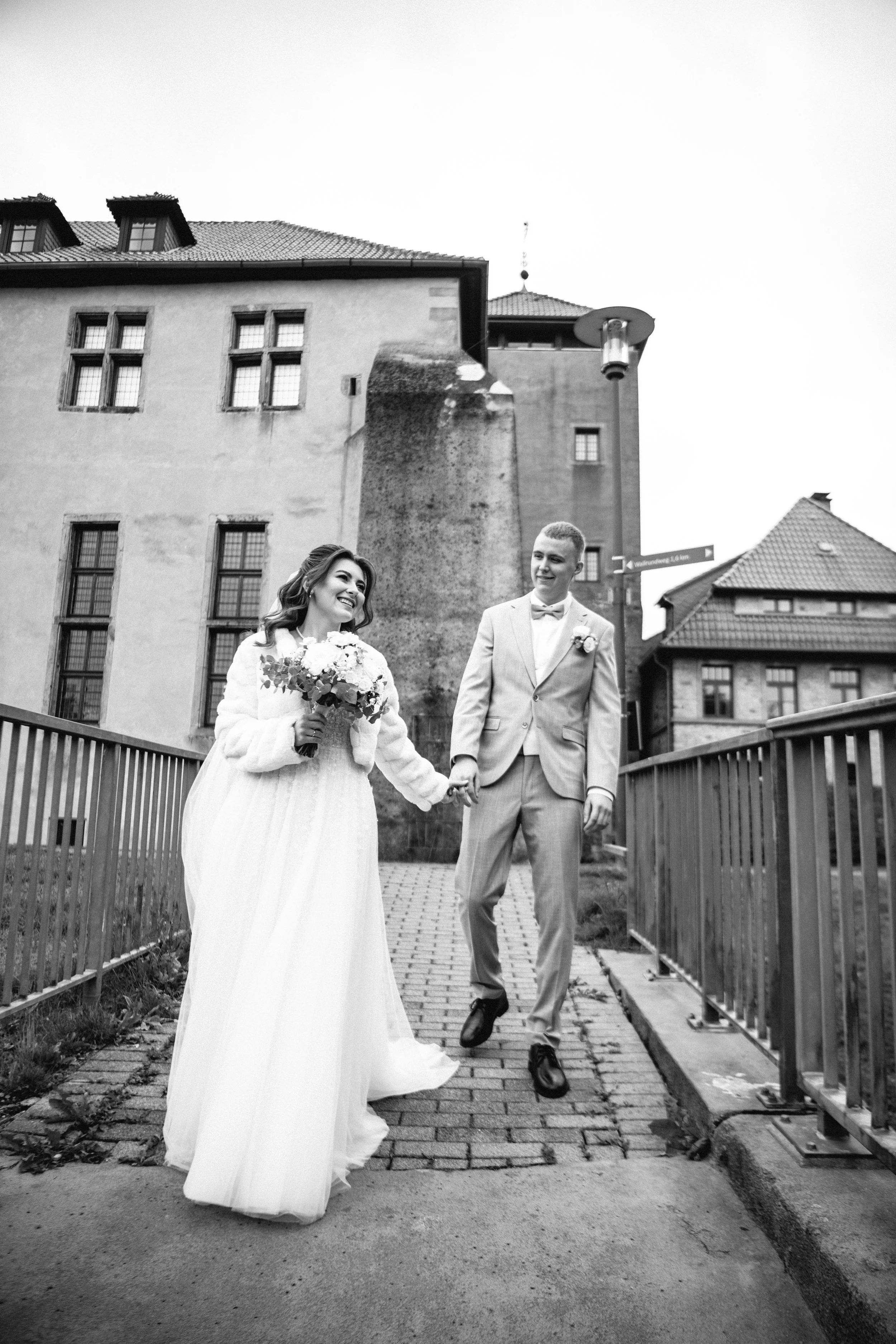 Black and white photo of a happy bride and groom holding hands while walking on a brick pathway outdoors.