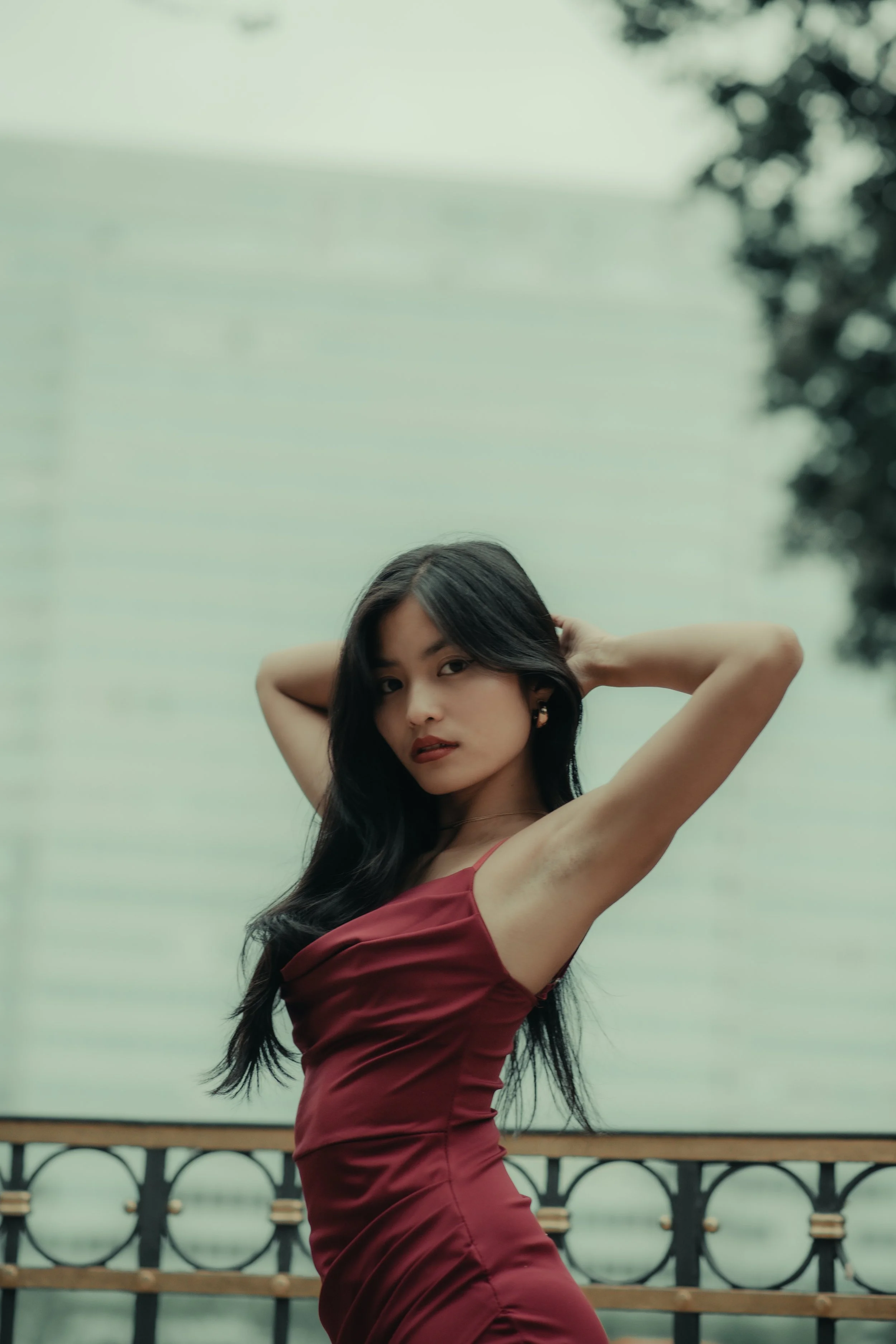 A young woman with long black hair wearing a fitted red sleeveless dress standing outdoors with a blurred background showing trees and a building.