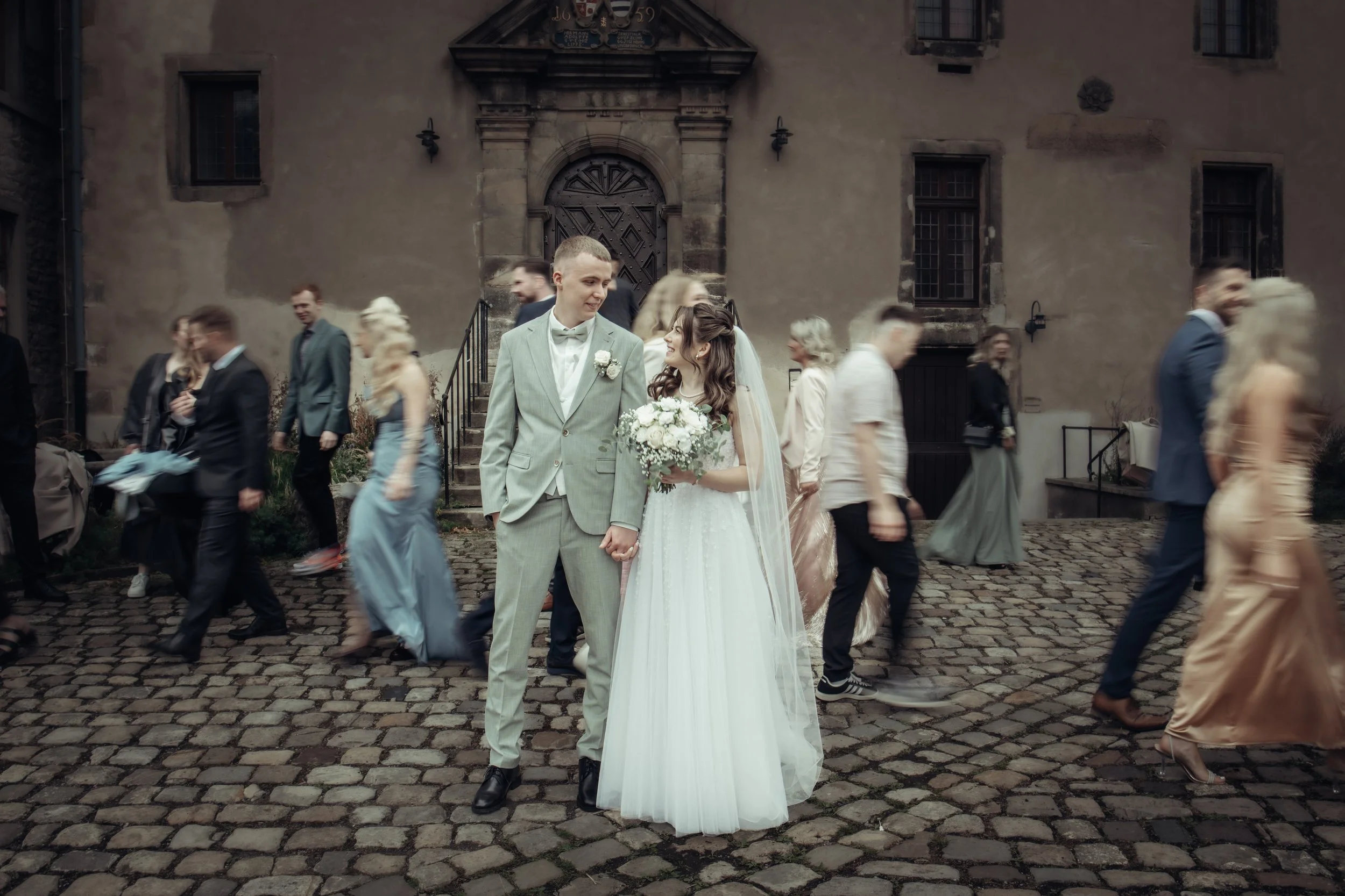 A bride and groom holding hands and looking at each other at their wedding celebration outdoors, with wedding guests walking in the background on a cobblestone street.