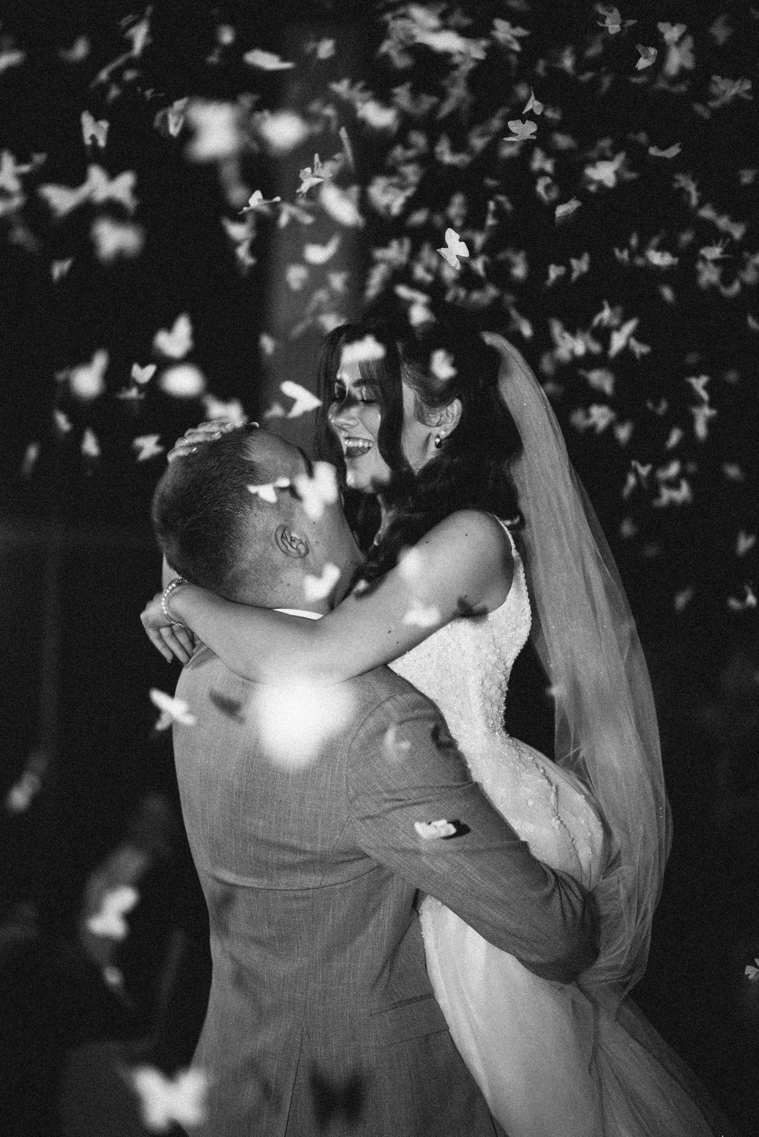A black and white photo of a bride and groom dancing, with falling confetti around them, at their wedding celebration.