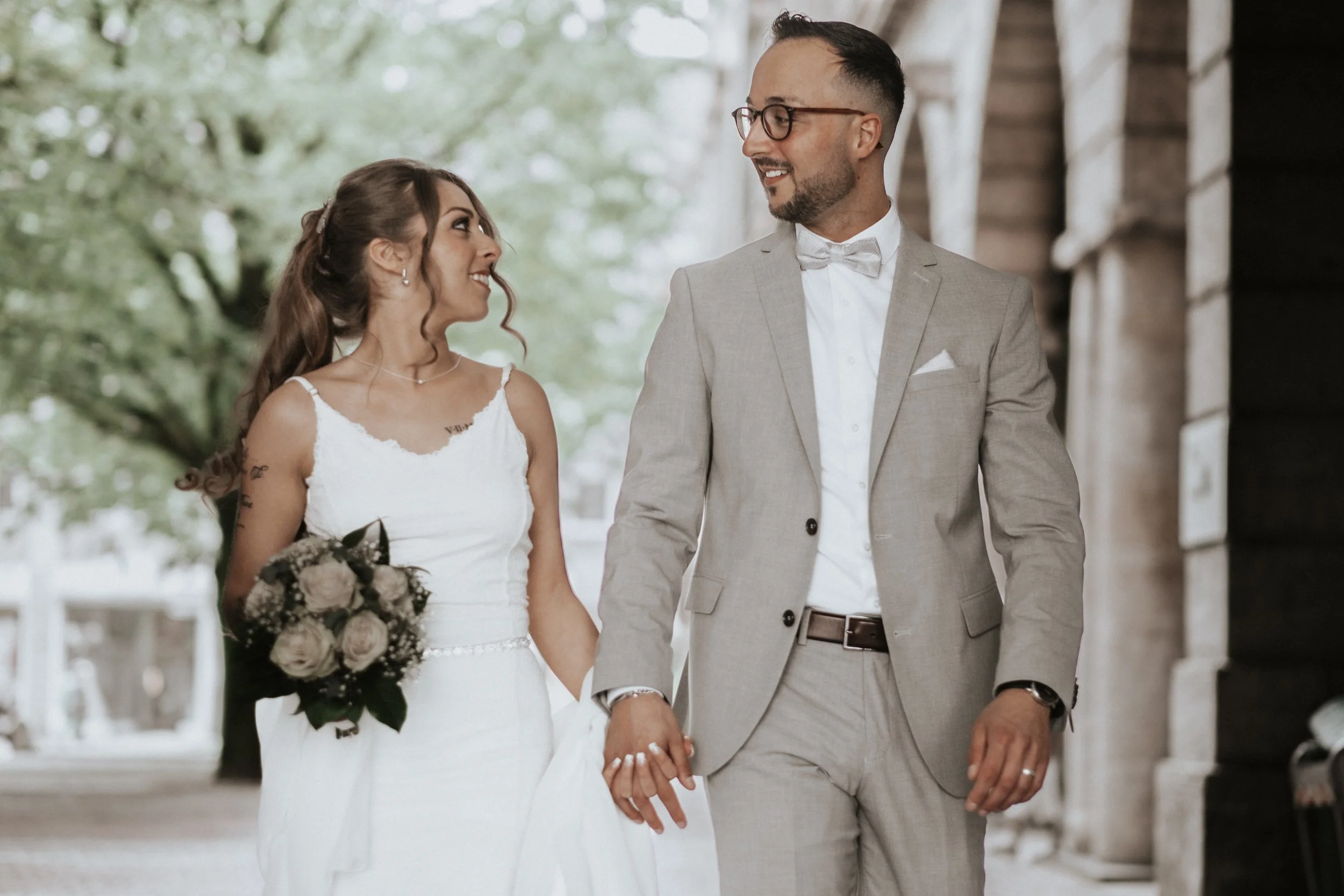 A couple walking hand in hand on their wedding day, with the bride in a white dress holding a bouquet of flowers and the groom in a beige suit with glasses.
