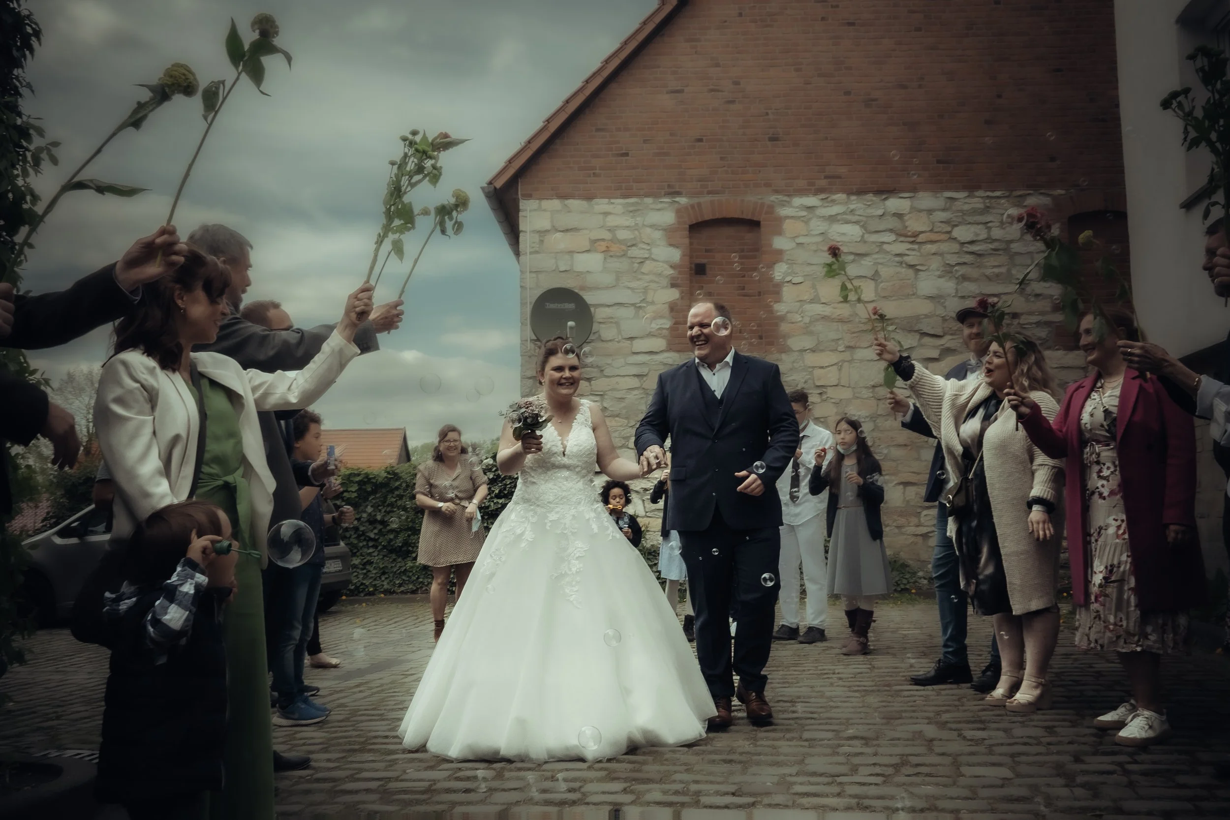 A newlywed couple walking hand in hand through a crowd celebrating their wedding outdoors, with guests holding flowers and bubbles.