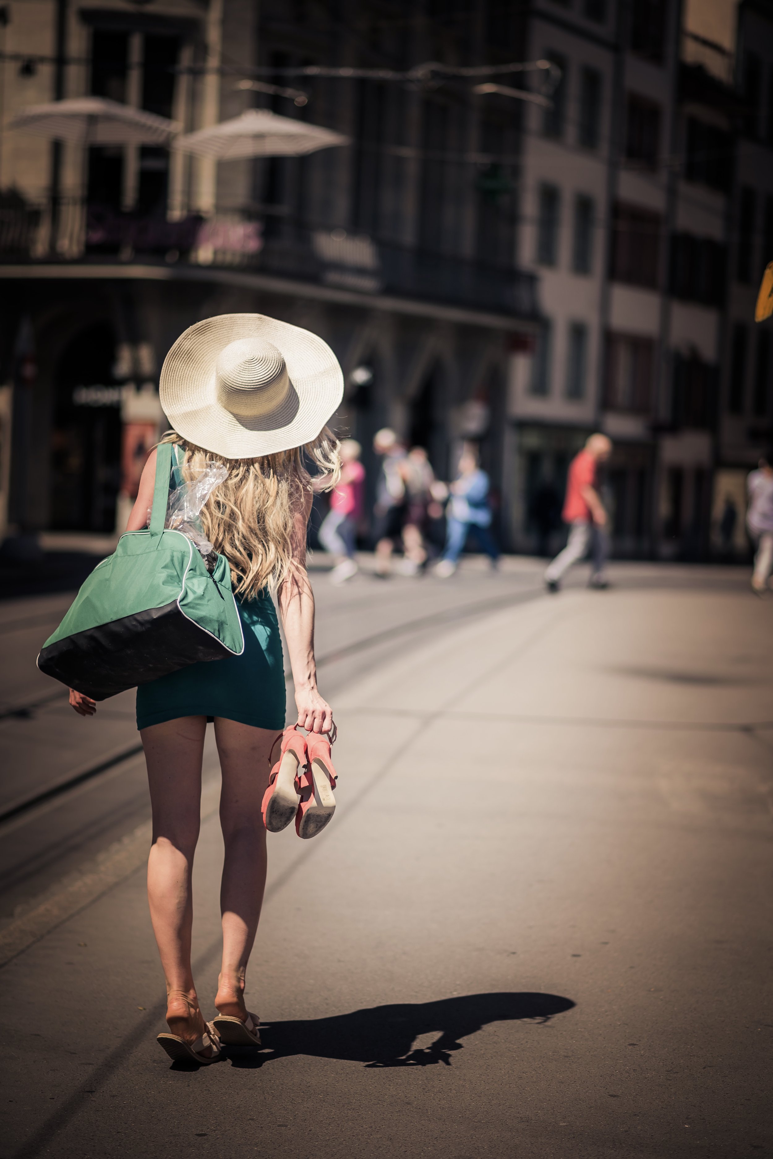 A woman walking on a city street carrying a green and black bag and a pair of pink shoes, wearing a large sun hat and a teal dress, with a shadow cast on the pavement and pedestrians in the background.