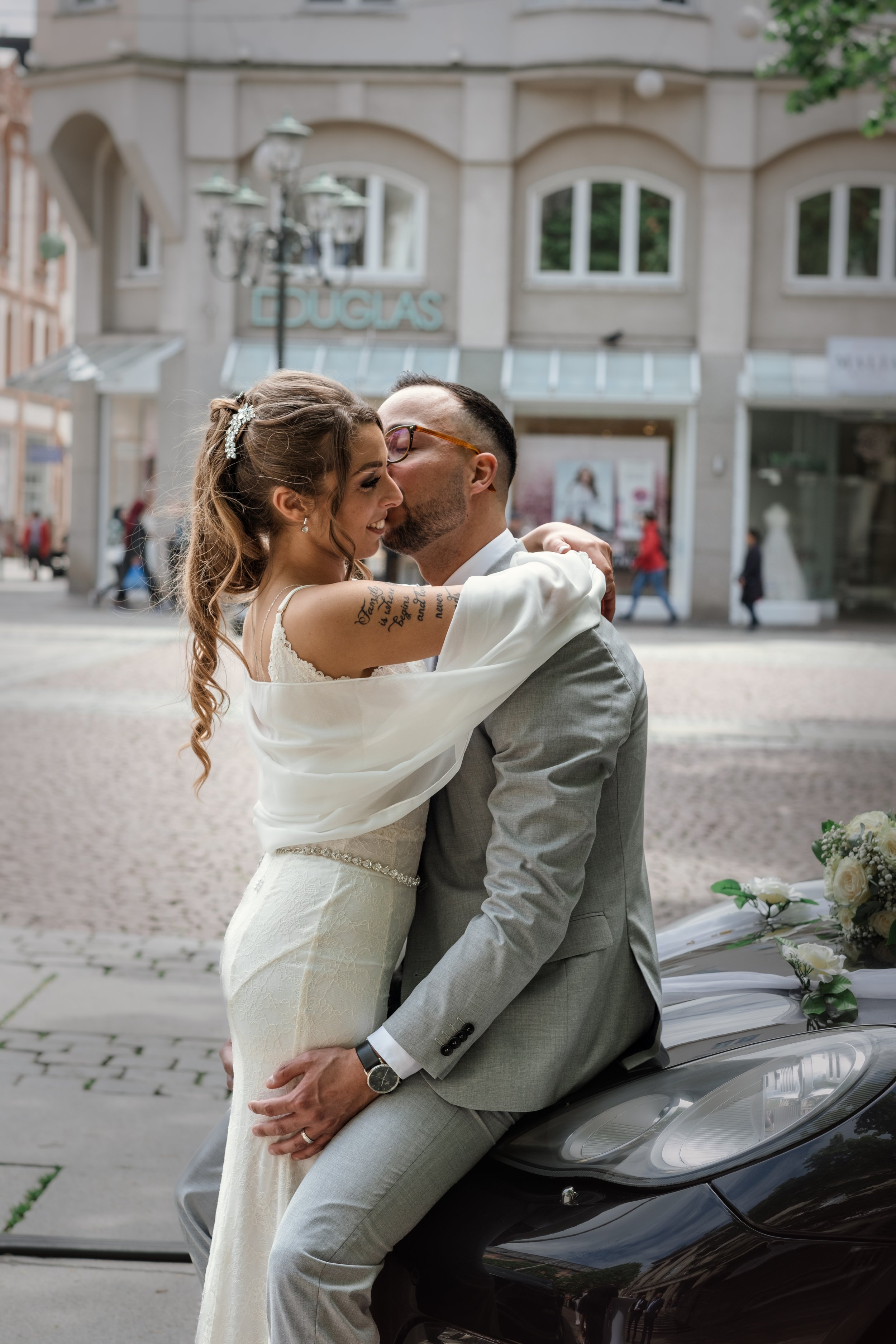 A newlywed couple embraces on a city street in front of a car decorated with flowers, smiling at each other as the bride and groom share a kiss.