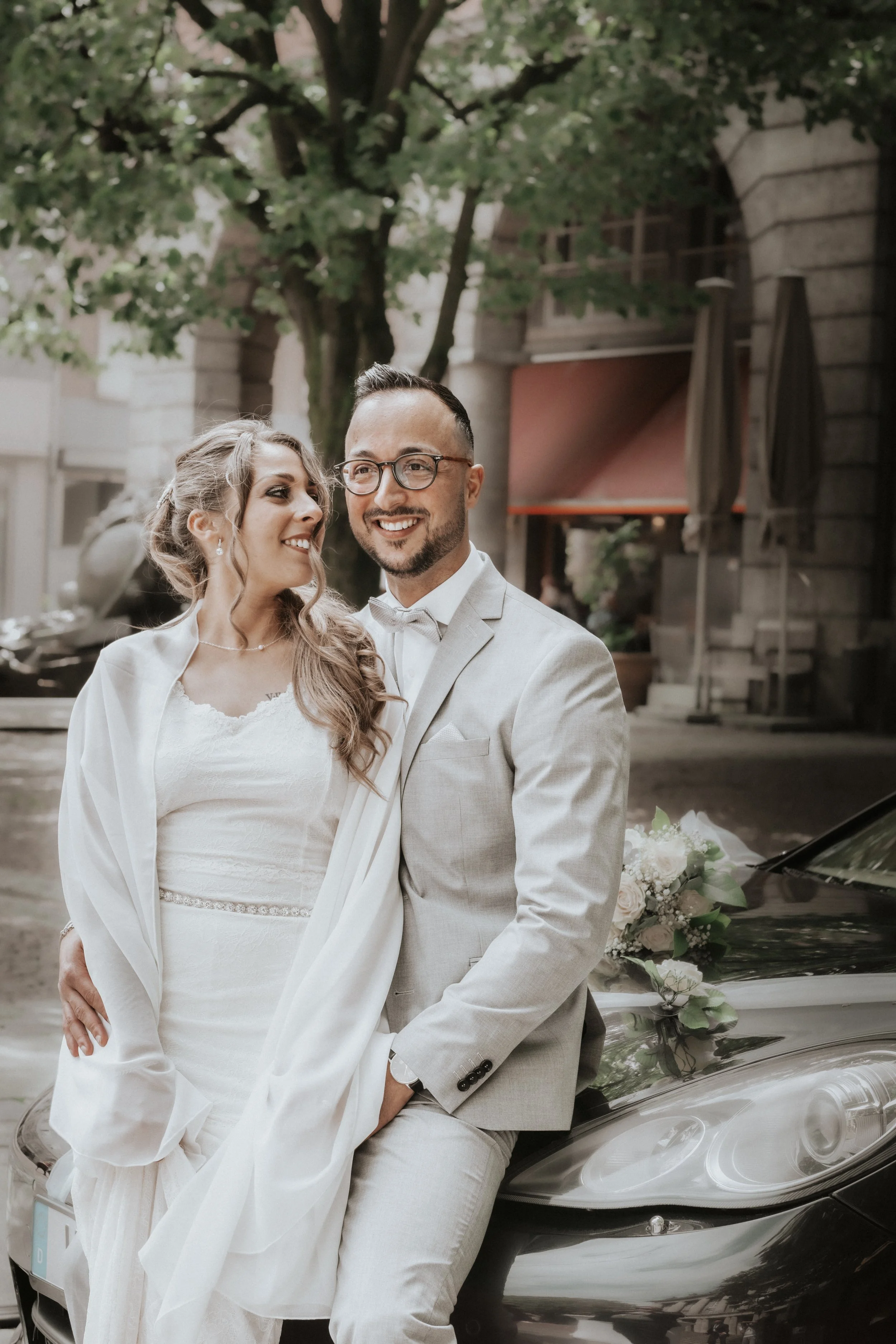 A newlywed couple dressed in white and gray wedding attire, sitting on the hood of a black car decorated with flowers, outdoors surrounded by trees.
