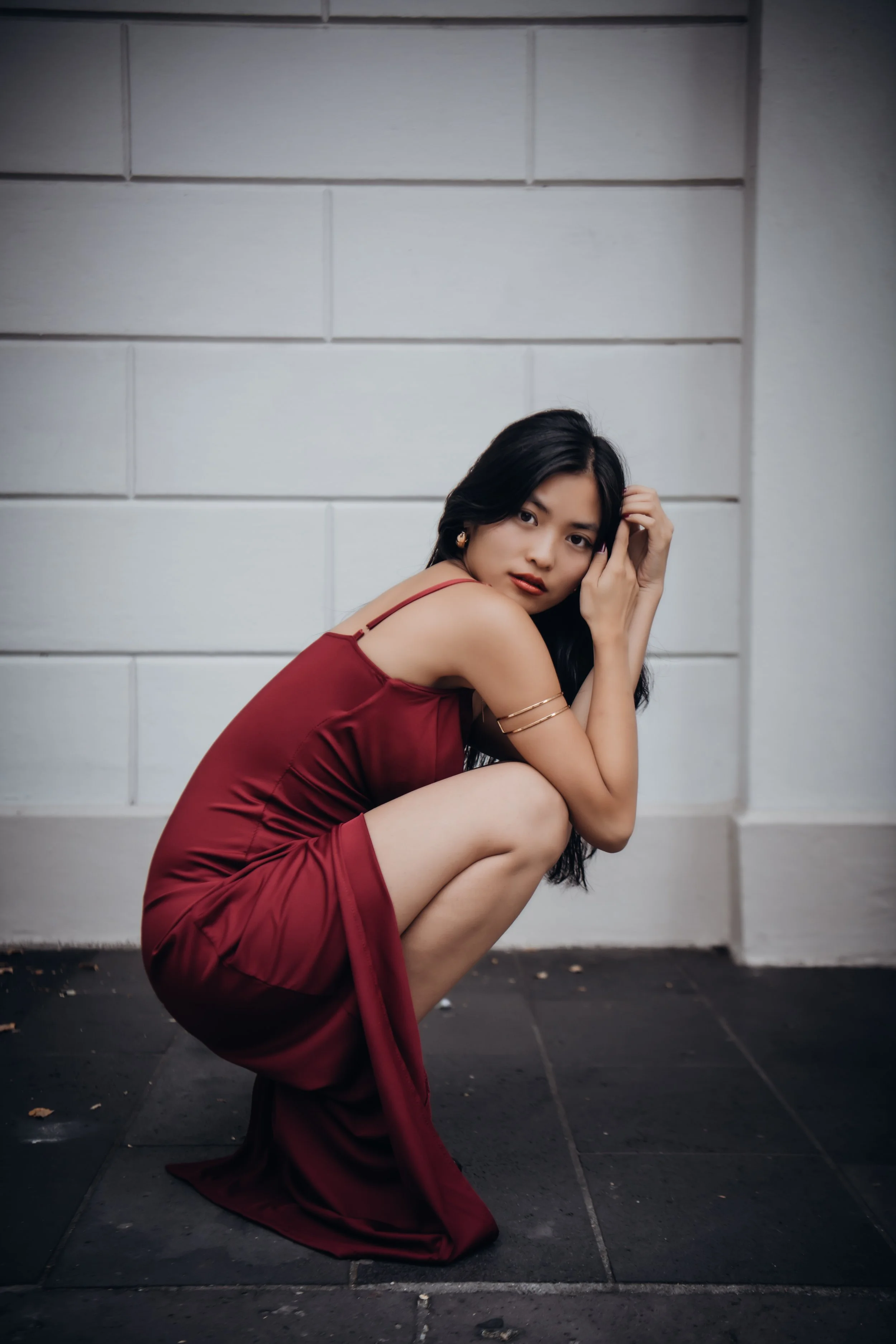 A woman in a burgundy dress crouches on the ground against a white brick wall, looking at the camera with her hand near her face.