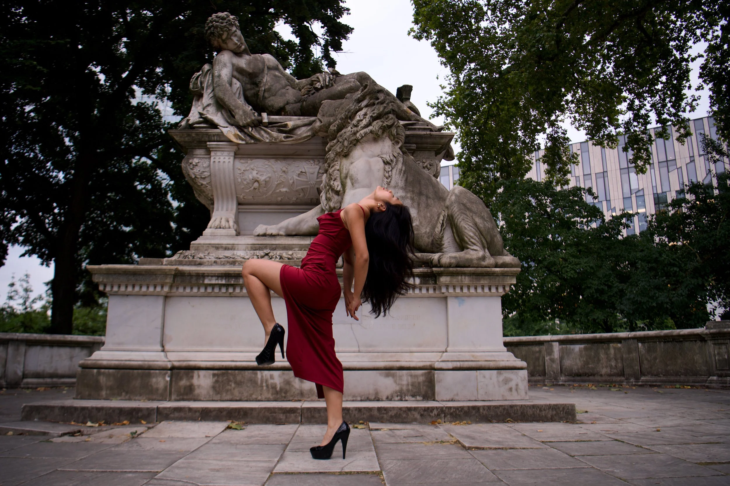 A woman in a red dress and black high heels posing in front of a large stone sculpture of a lion and a person lying on top of it, in a park with trees and a building in the background.
