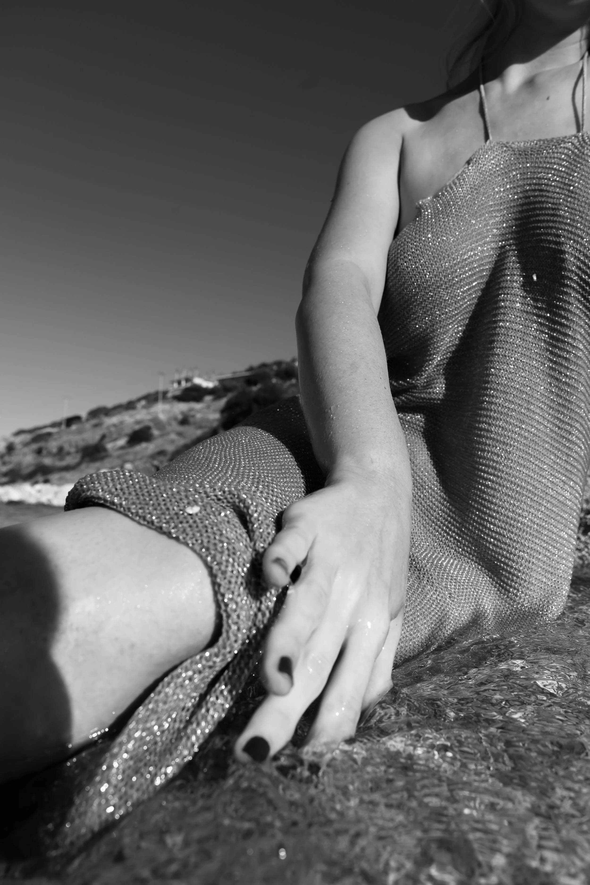 A close-up black and white photo of a woman in a glittery dress lying on the beach with her arm extended and hand touching the water, with a hilly landscape in the background.