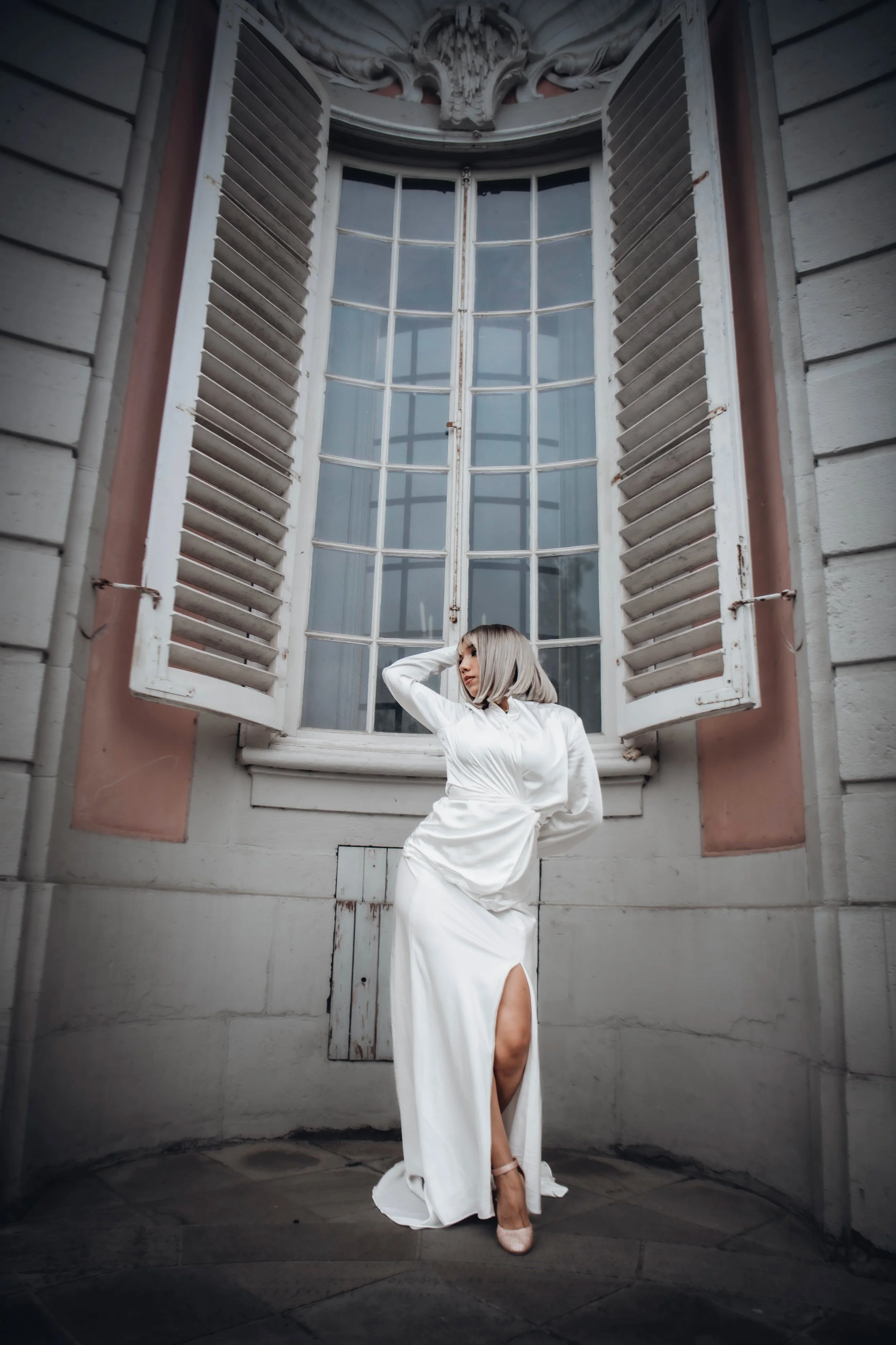 A woman in a white gown standing in front of a large window with open shutters in an ornate building.