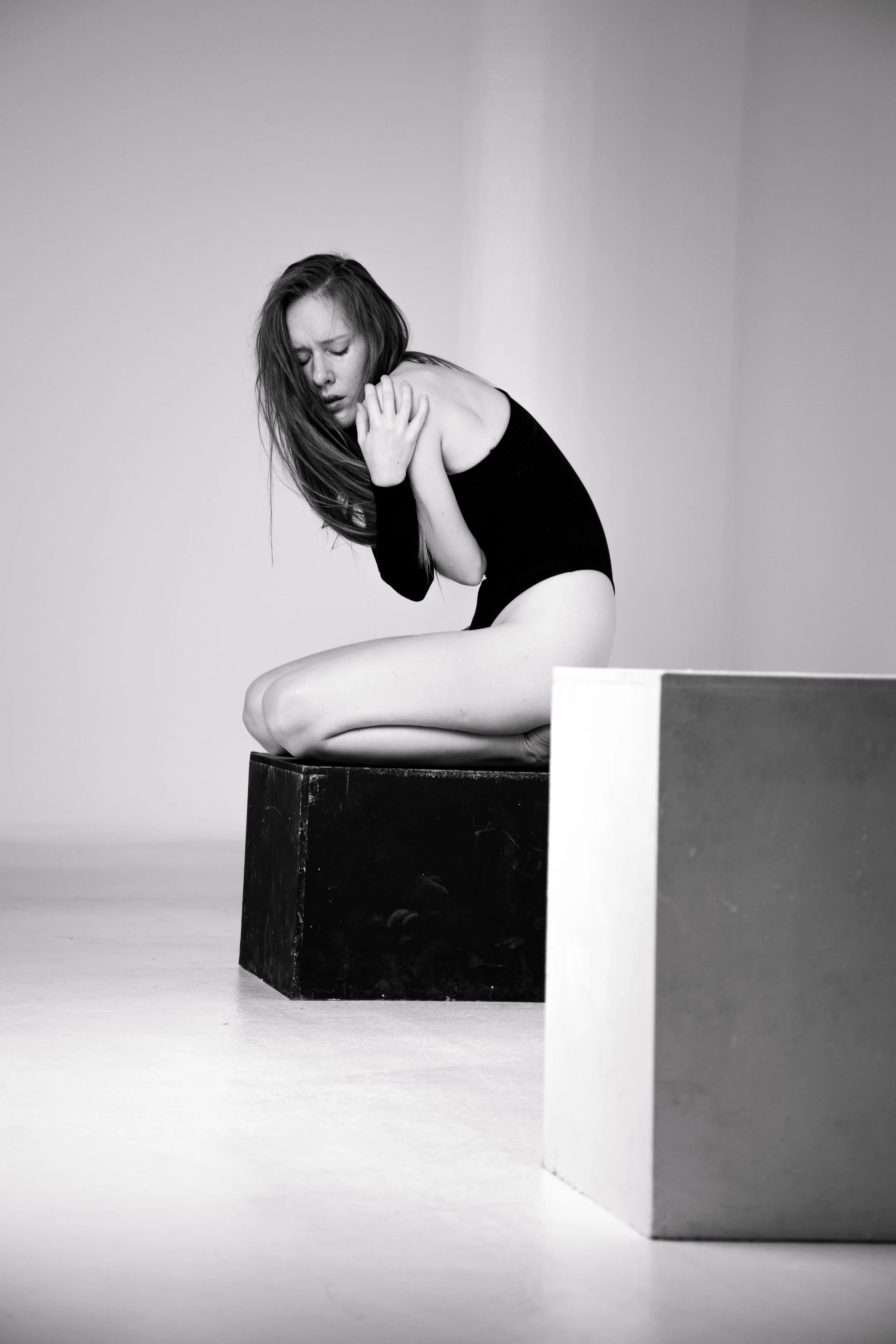 A black and white photo of a woman sitting on a dark cube, her head bowed, with her hand on her shoulder in a pensive pose. She wears a dark sleeveless top and her long hair falls over her face. The background is plain and minimalistic.