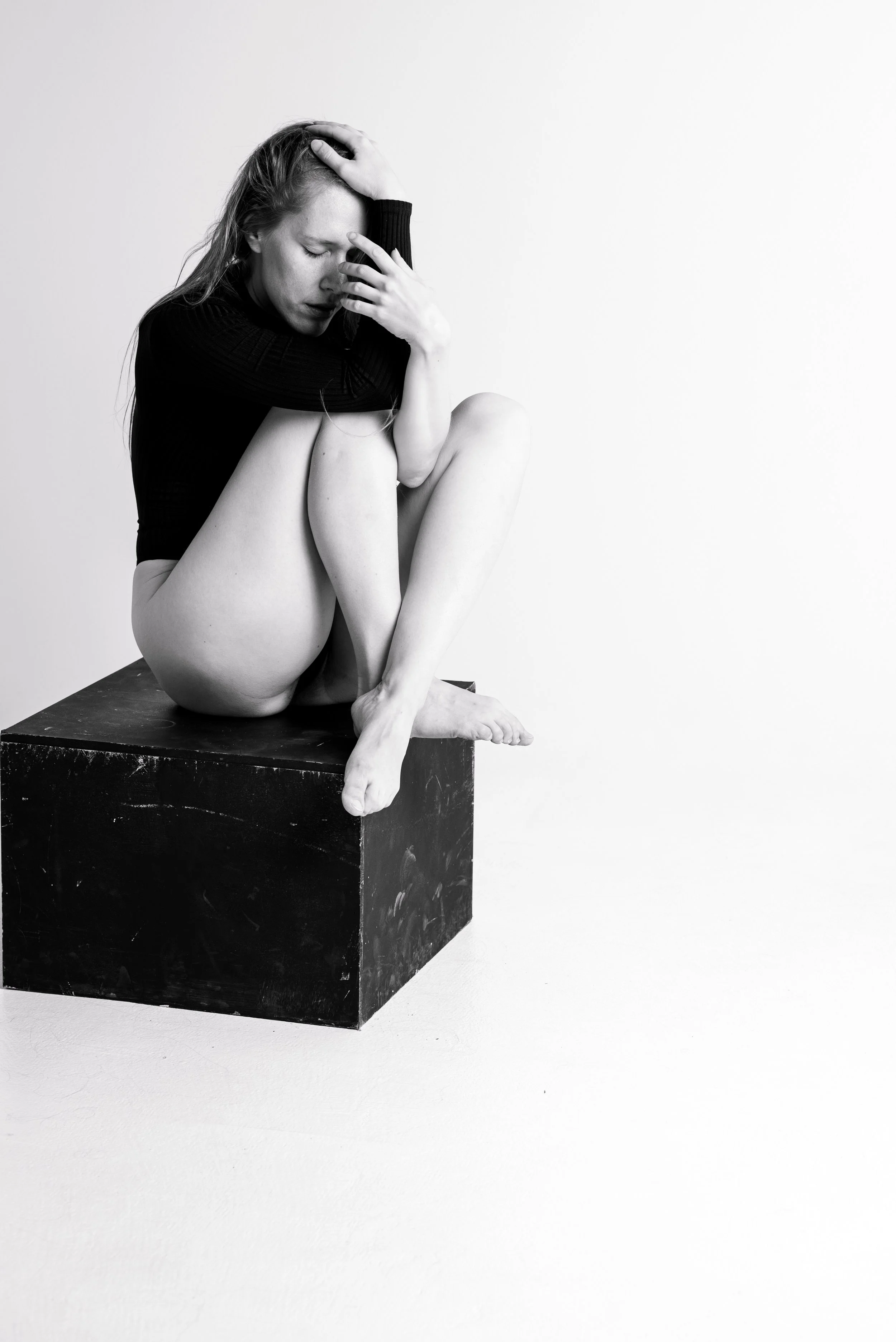 Black and white photo of a woman sitting on a box with her knees drawn up, her head resting in her hand, and an expression of distress or deep thought.