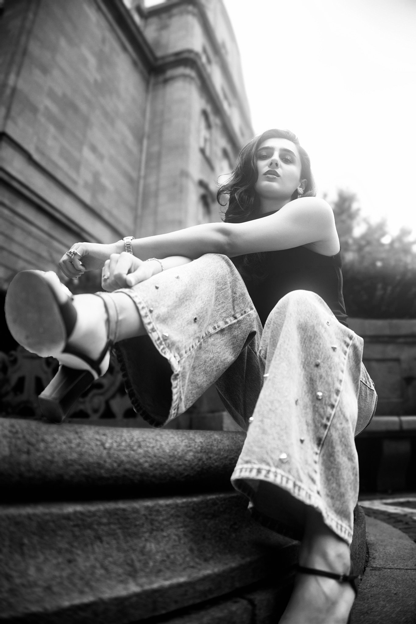 A black and white photo of a woman sitting on outdoor steps, looking at the camera with a confident expression. She's wearing a sleeveless top, jeans, and a watch, with her legs crossed and one arm resting on her knee. The background shows a stone bu