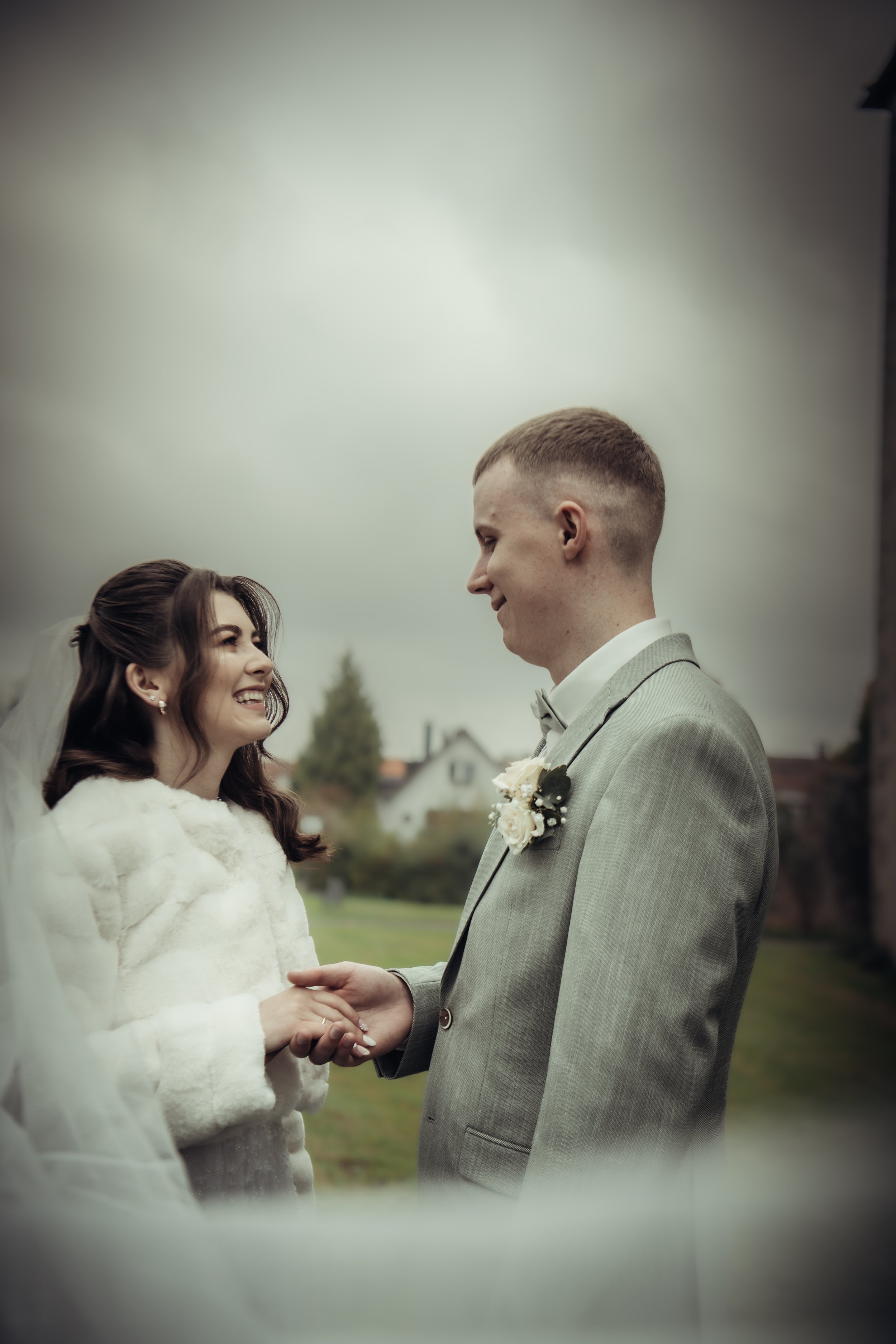 A bride and groom holding hands and smiling at each other outdoors during a wedding ceremony on a cloudy day.