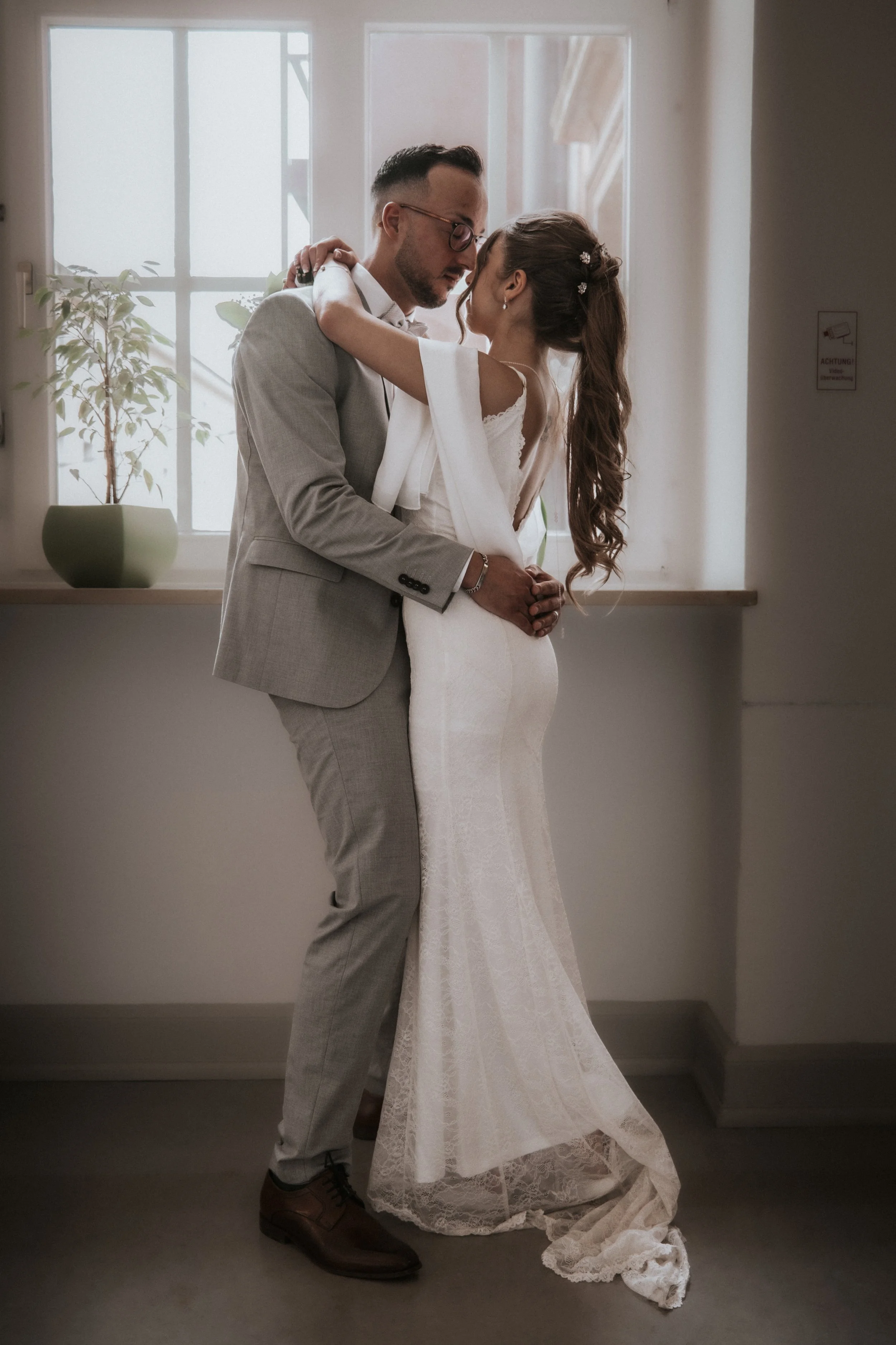 A bride and groom sharing an intimate dance indoors near a window, with the groom in a light gray suit and the bride in a white wedding gown.