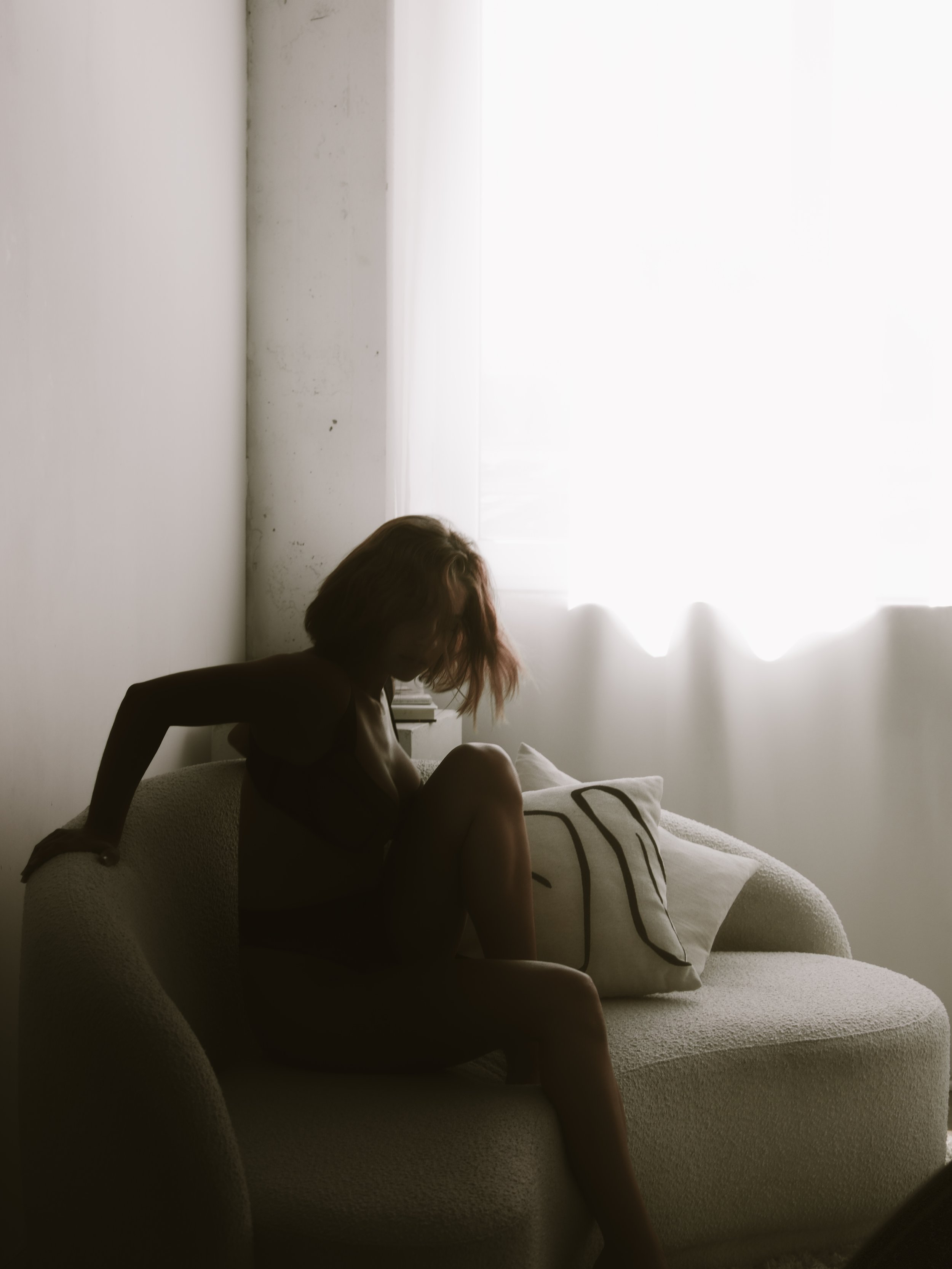 A person sitting on a light-colored sofa near a window with bright white curtains, their head bowed and hair obscuring their face, in a room with white walls.