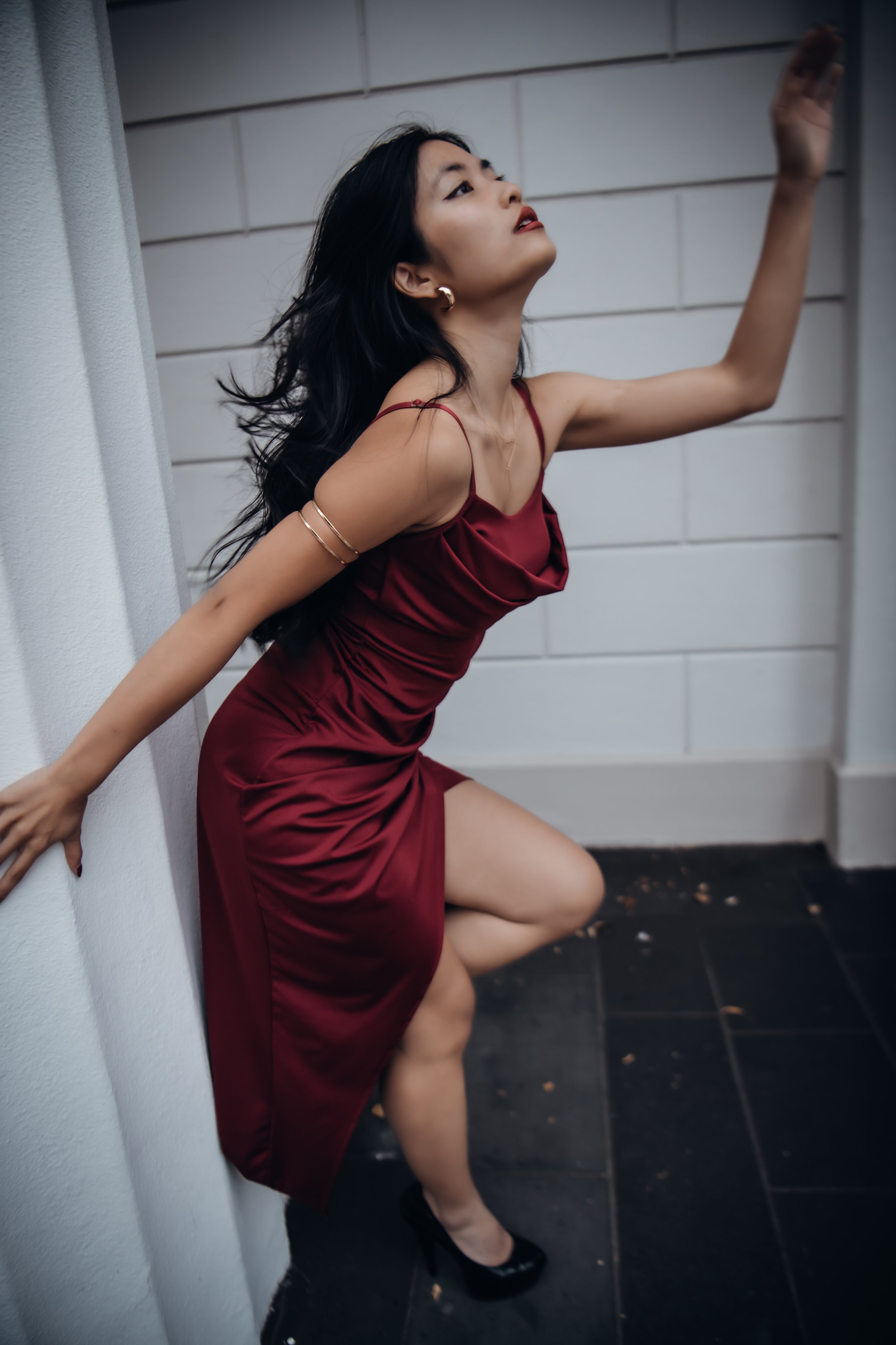 A woman with long black hair wearing a red satin dress and black high heels, leaning against a white wall with one hand on the wall and the other raised, with a serious expression, against a background of white walls and a black tiled floor.