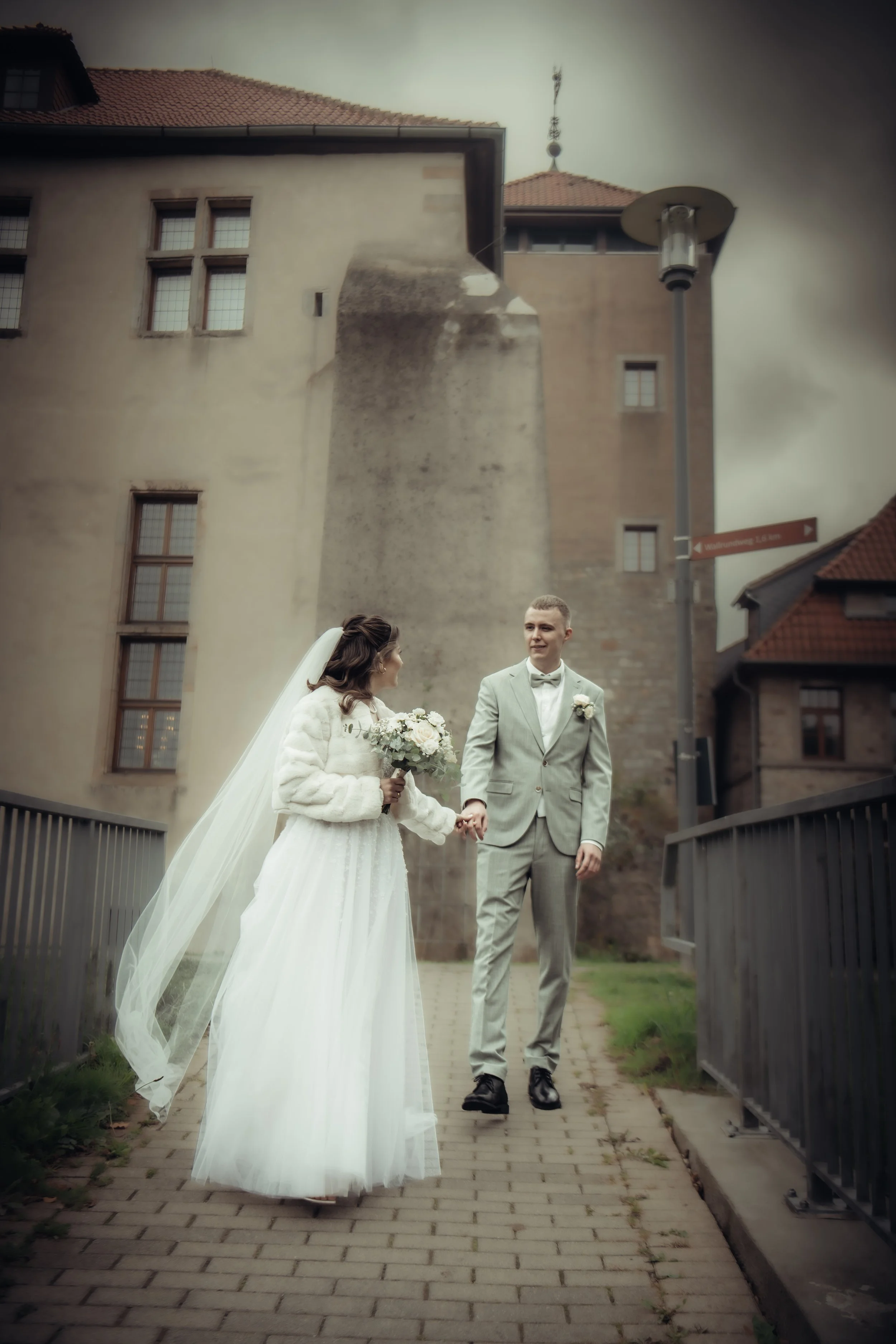 Bride and groom holding hands on a cobblestone pathway in front of a historic building, with the bride in a white wedding dress and the groom in a gray suit.