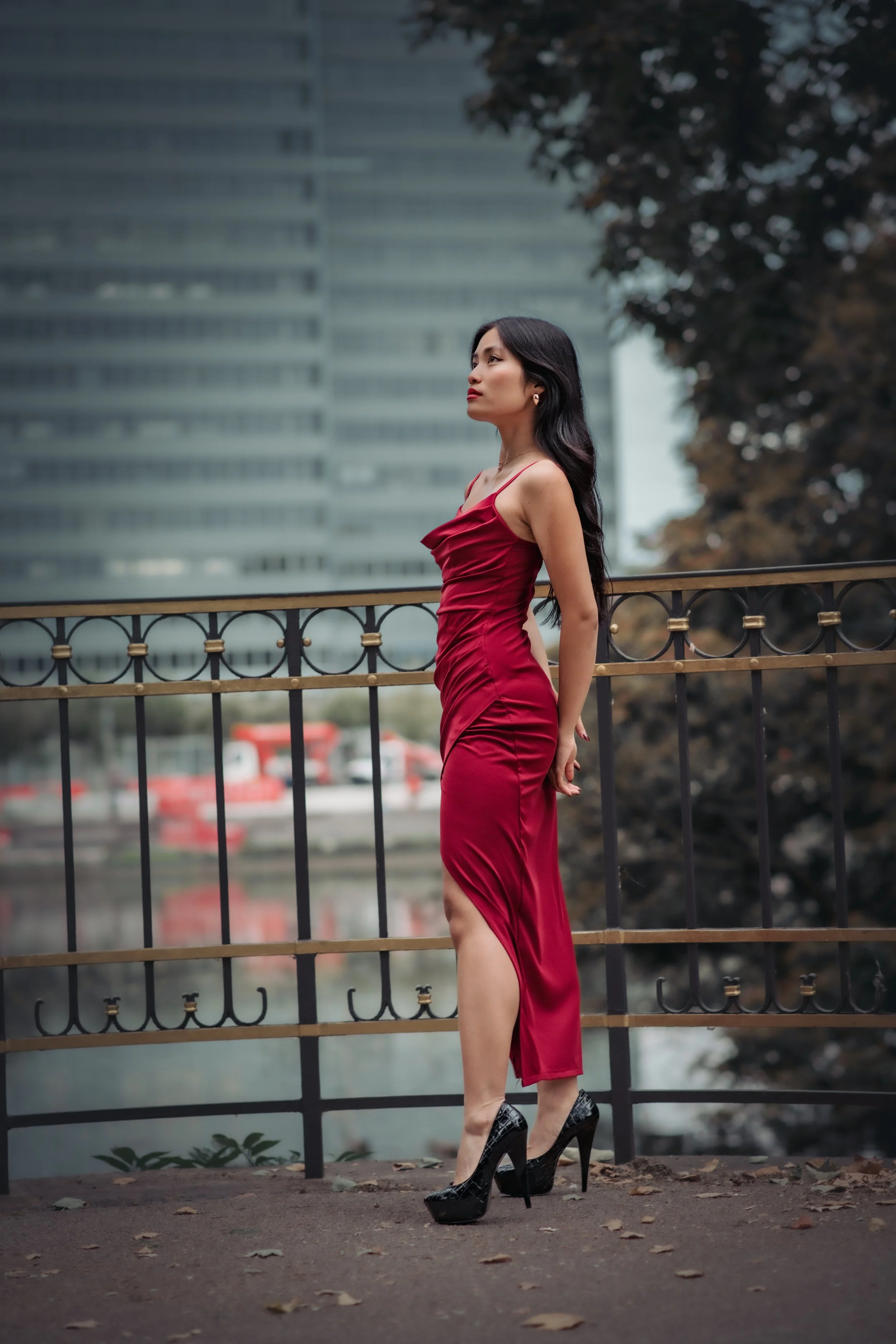 A woman in a red satin slip dress and black high heels standing on a bridge with a city skyline and trees in the background.