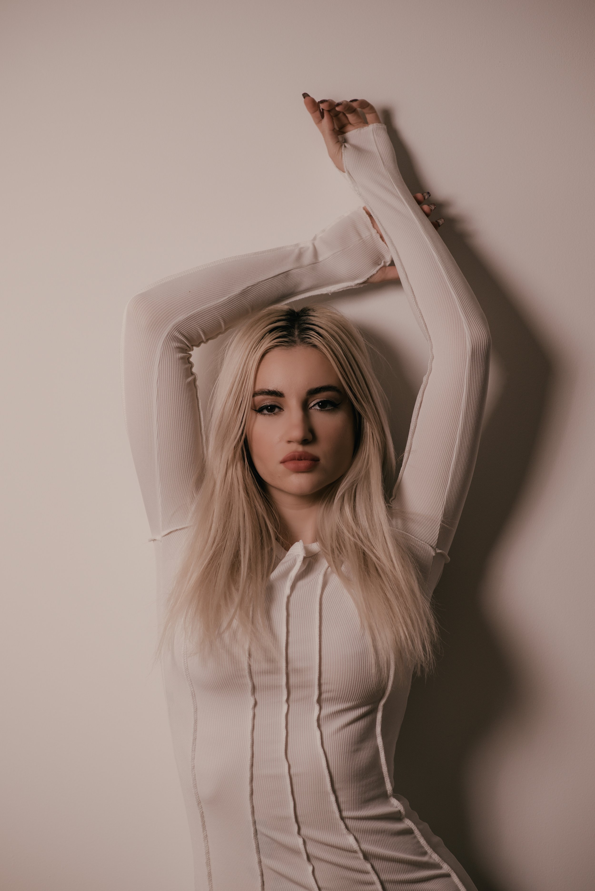 A young woman with long blonde hair posing with arms raised above her head, wearing a beige, long-sleeved, fitted top, standing against a plain neutral-colored wall.