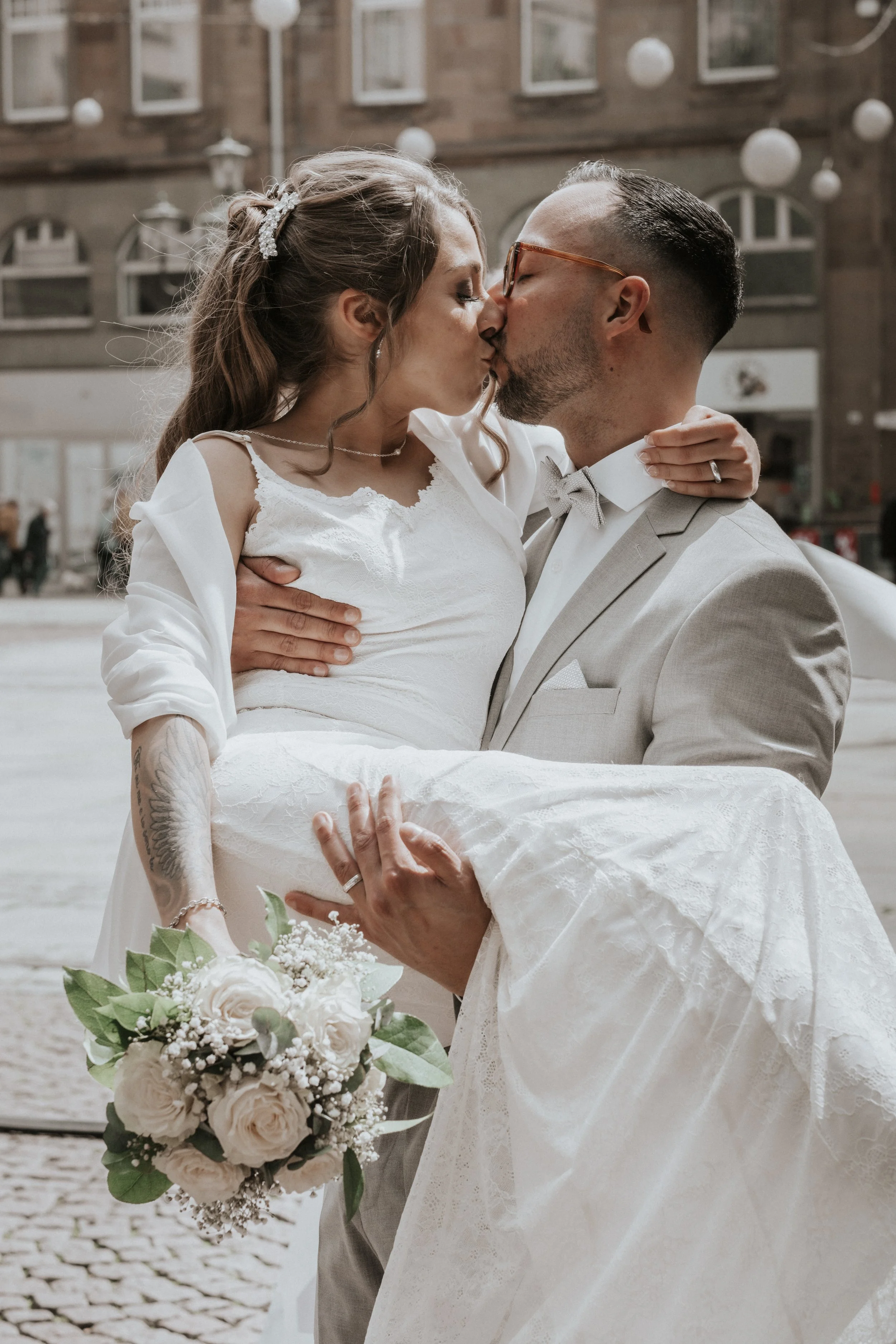 A newlywed couple shares a kiss outside, with the groom lifting the bride. The bride holds a bouquet of white roses and greenery, and the bride and groom are dressed in wedding attire, with the groom in a light gray suit and the bride in a white lace