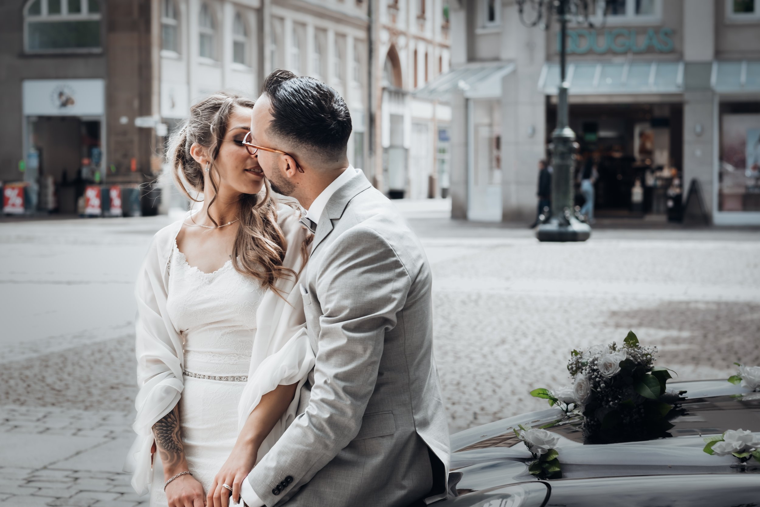 A newlywed couple sharing a kiss on a city street, with a wedding bouquet on a nearby car.