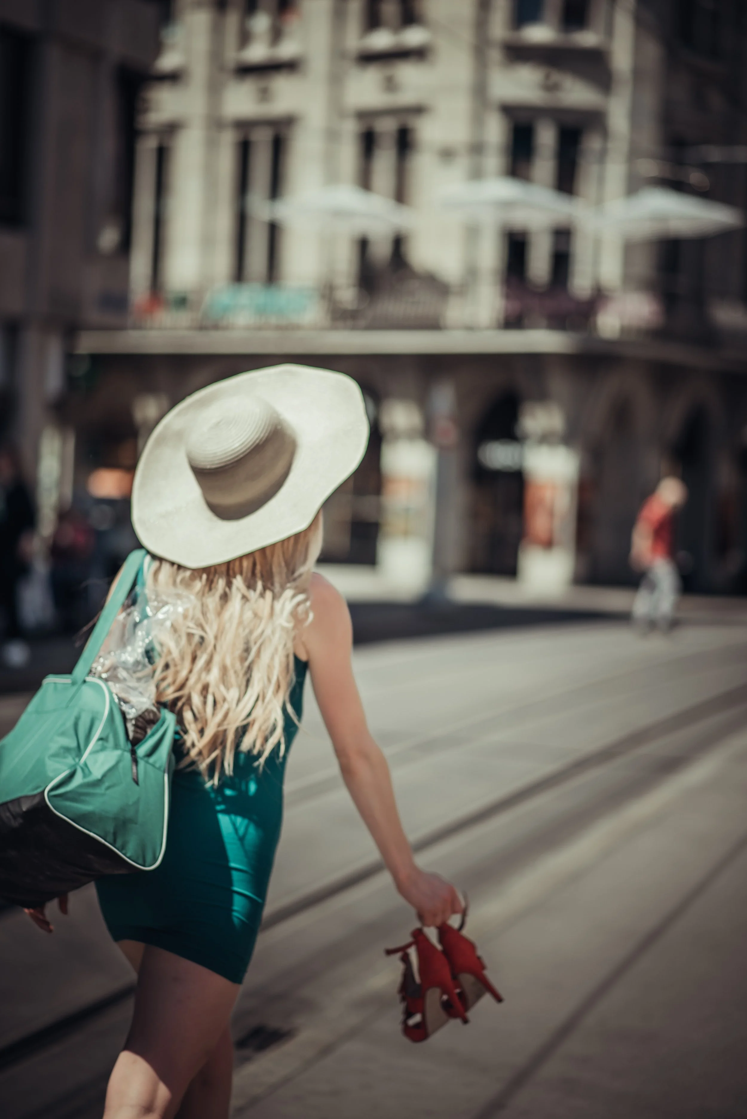 A woman with long blond hair wearing a large sun hat, teal dress, carrying a green and black bag, and holding red high heels, walking along tram tracks in an urban setting.