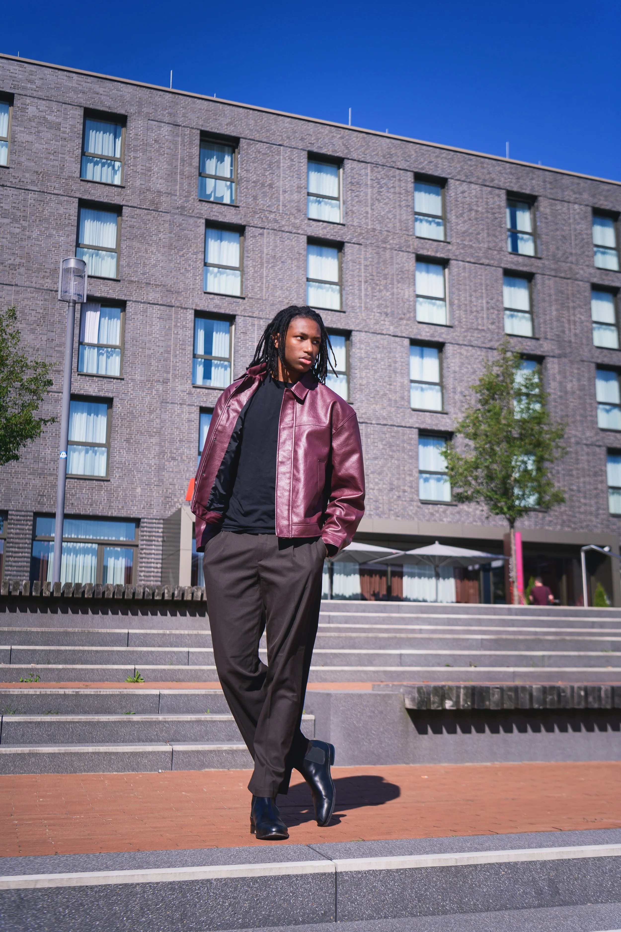 A woman in a black shirt, burgundy leather jacket, gray pants, and black ankle boots stands on steps outside a modern brick apartment building under a clear blue sky.