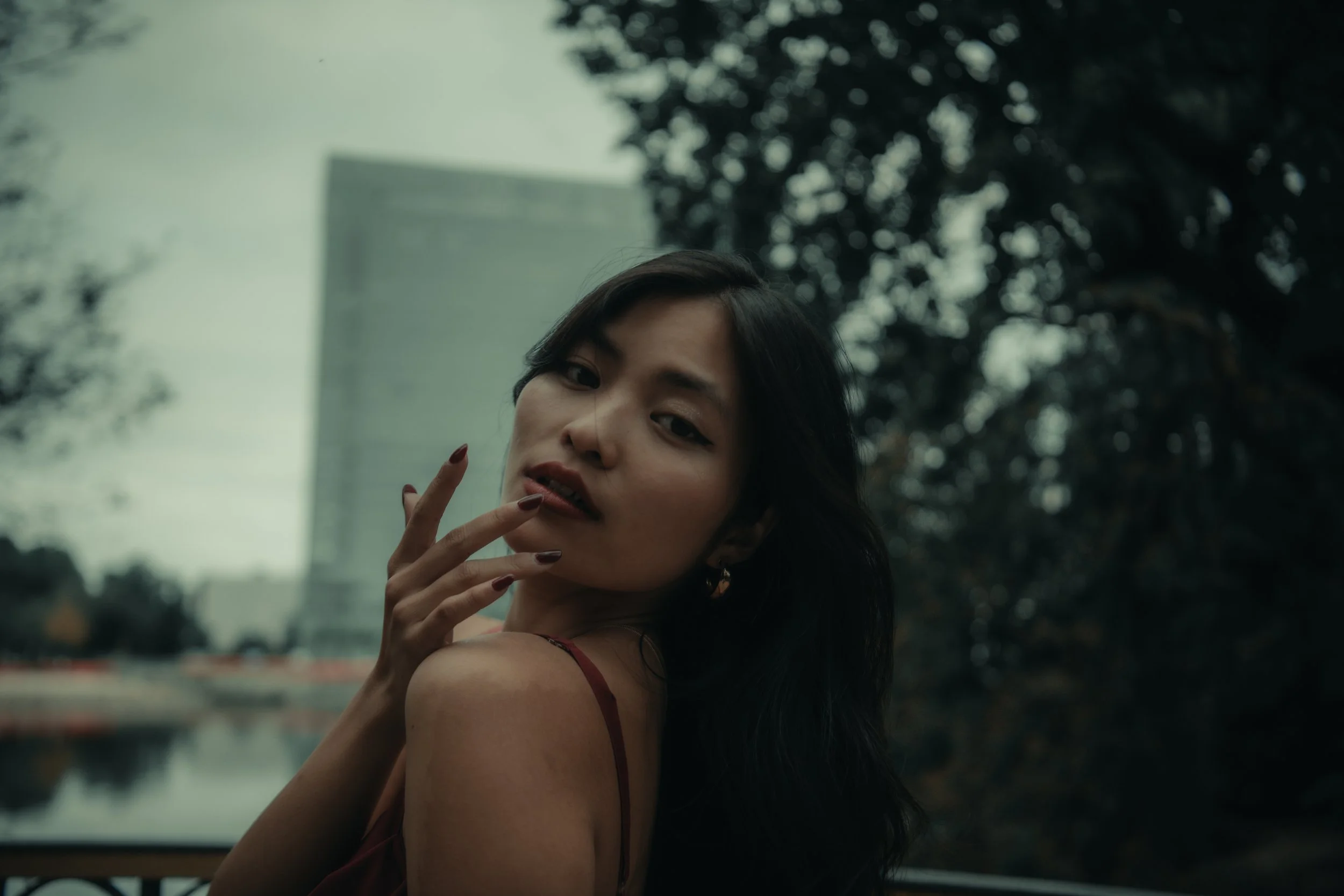 A woman with dark hair and red lipstick posing outdoors near a river with a cityscape and trees in the background.
