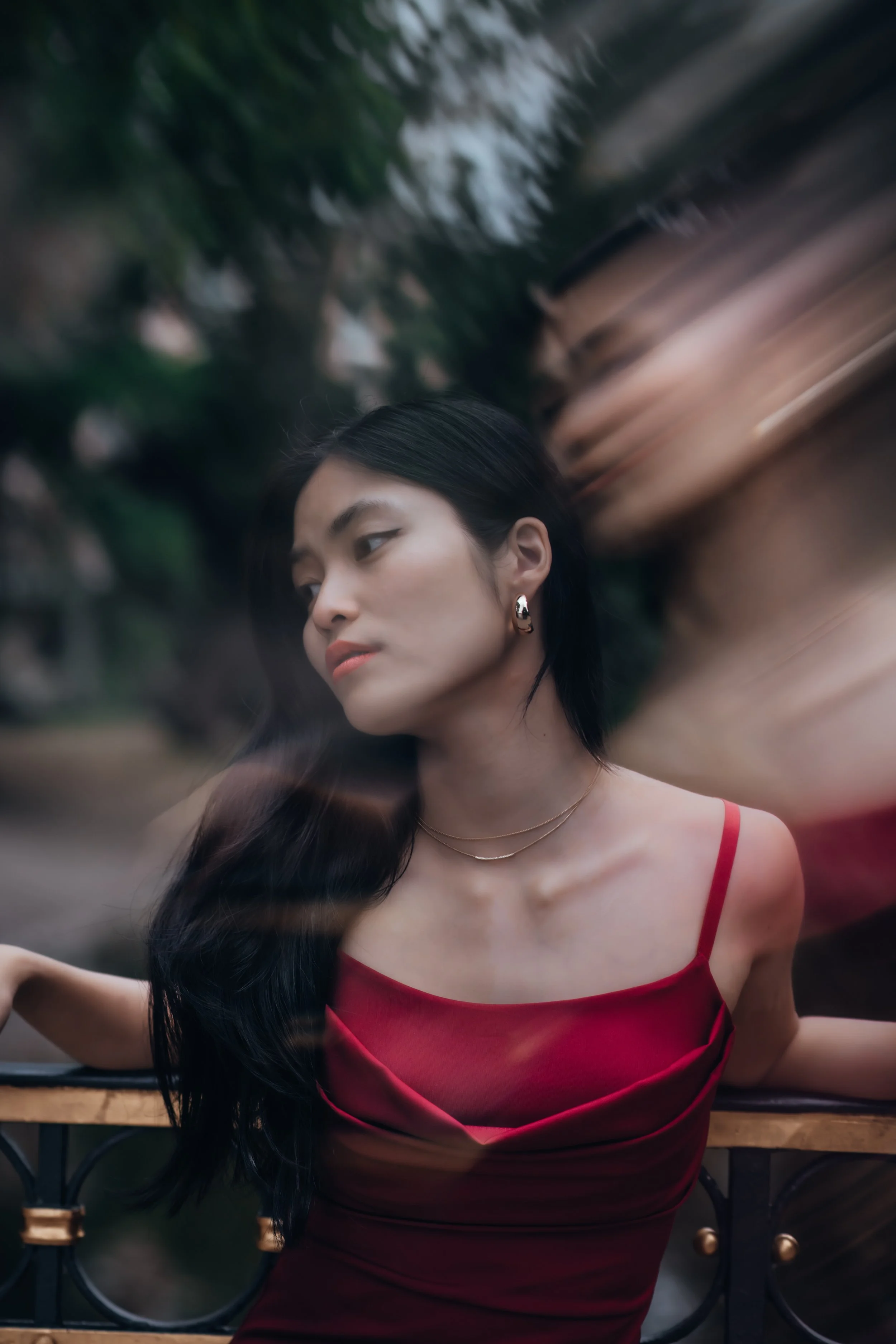 A woman in a red dress and earrings stands outside near a railing, with her eyes closed and dark hair flowing over her shoulder. A blurred figure appears behind her, creating a motion effect.