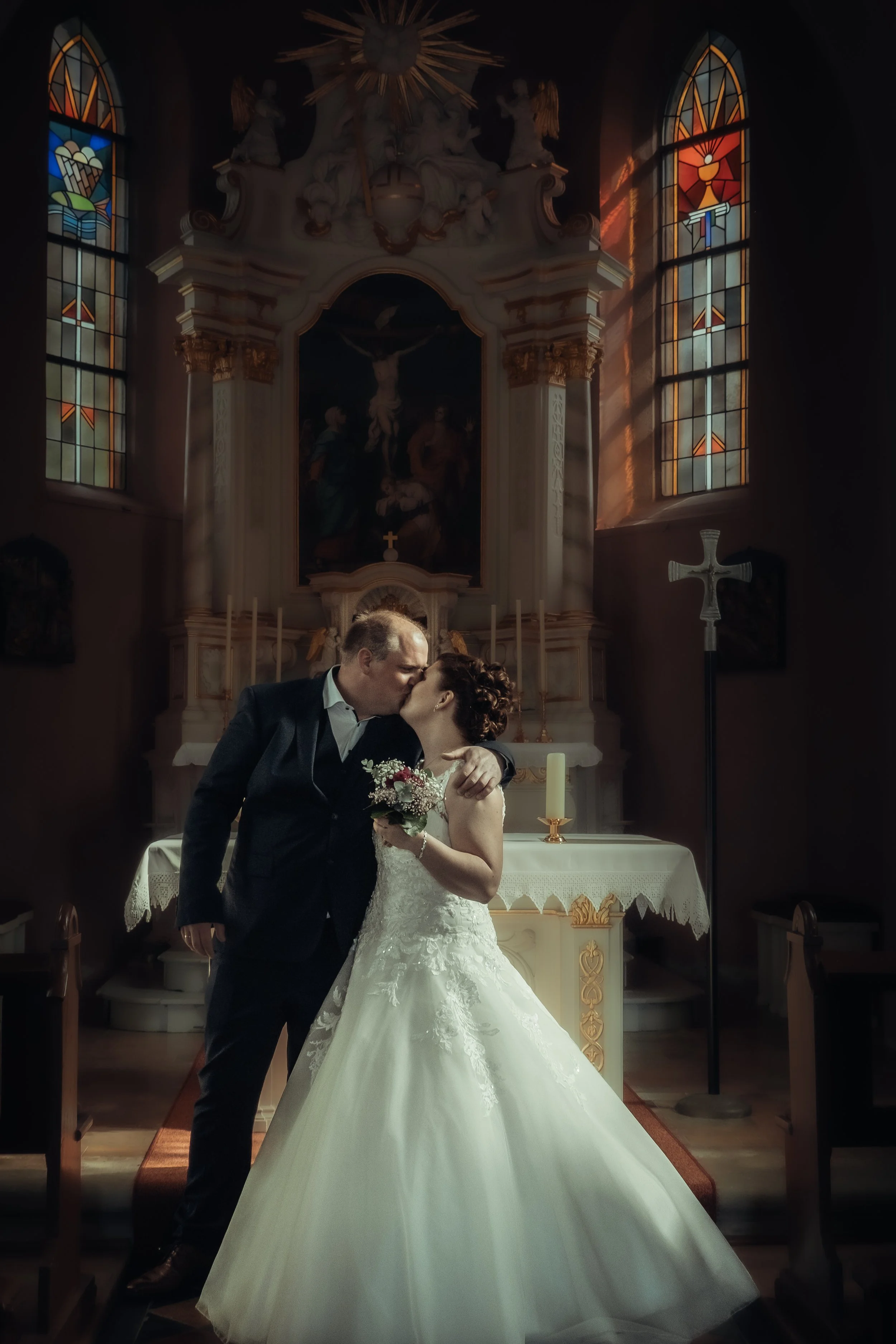 A bride and groom kissing inside a church altar during their wedding ceremony, with stained glass windows and religious symbols visible.