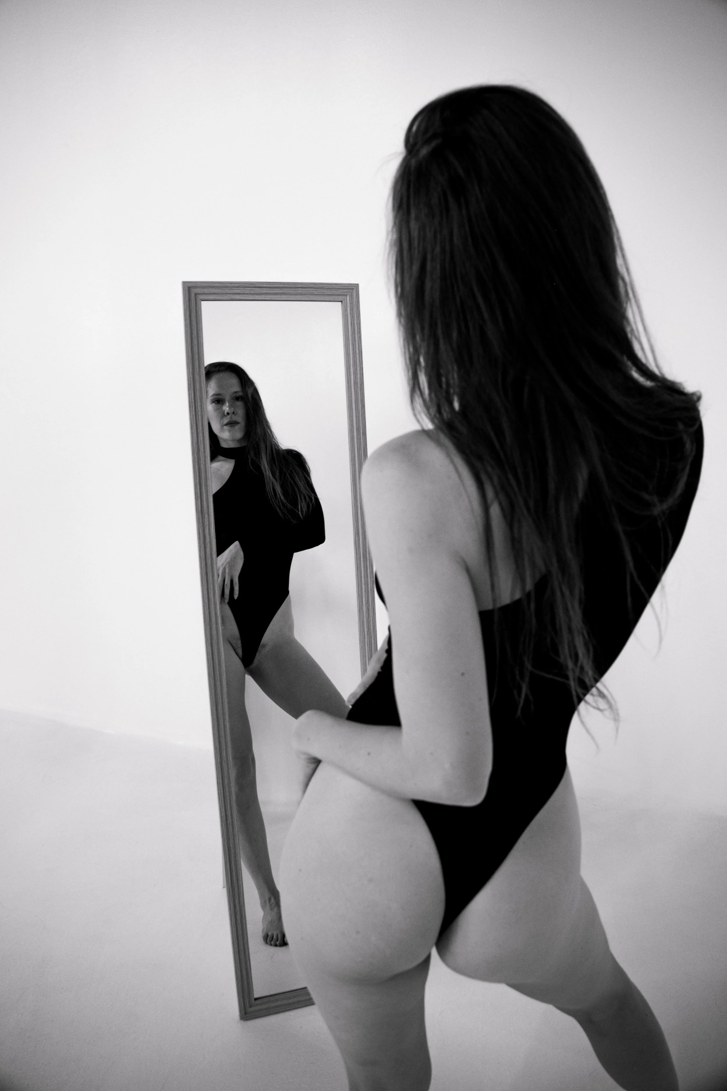 A woman with long hair wearing a black bodysuit, looking at her reflection in a mirror.