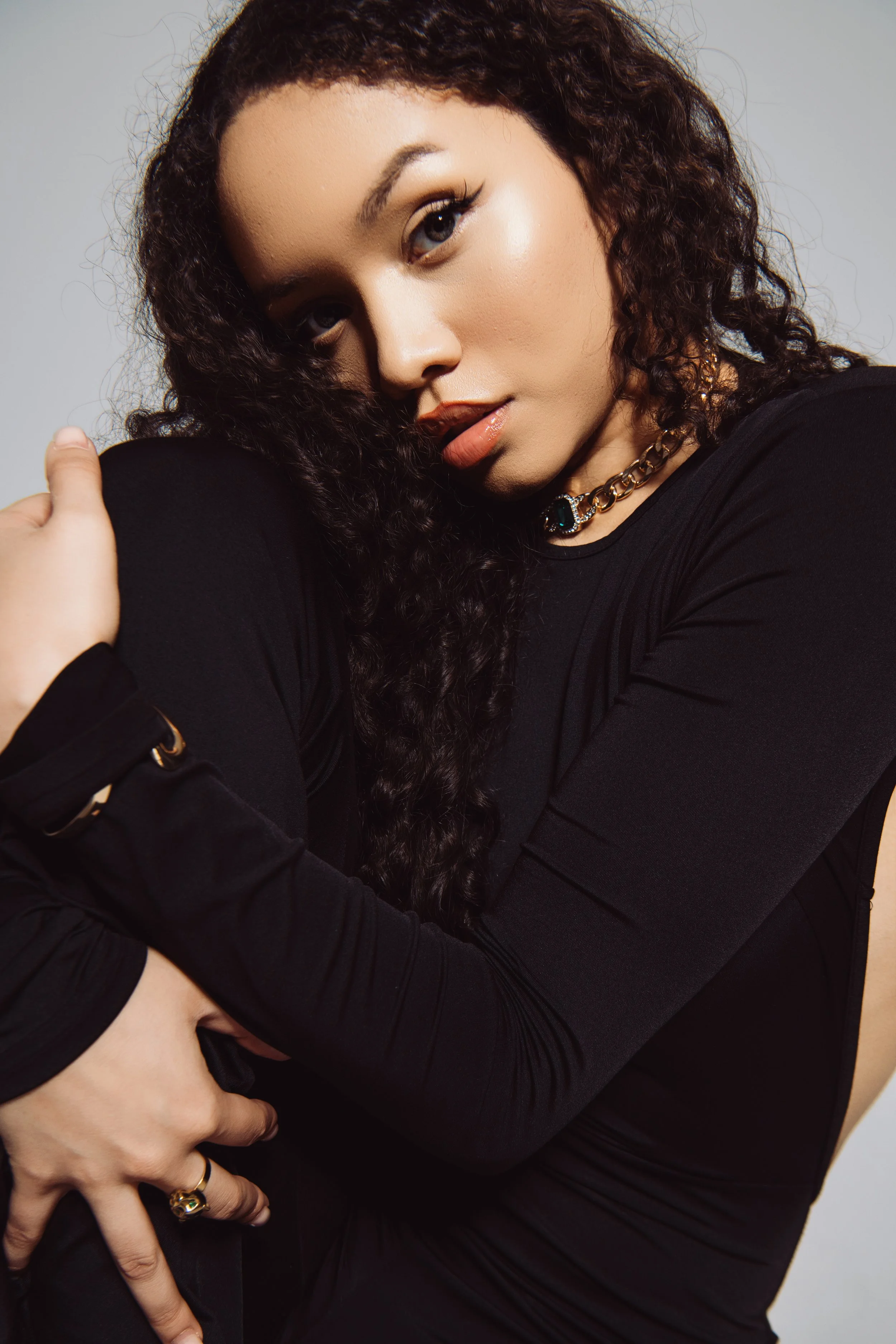 A young woman with curly dark hair, wearing a black long-sleeve top and gold jewelry, embraces herself with her arms crossed in front. She has a confident expression and is looking at the camera against a plain background.