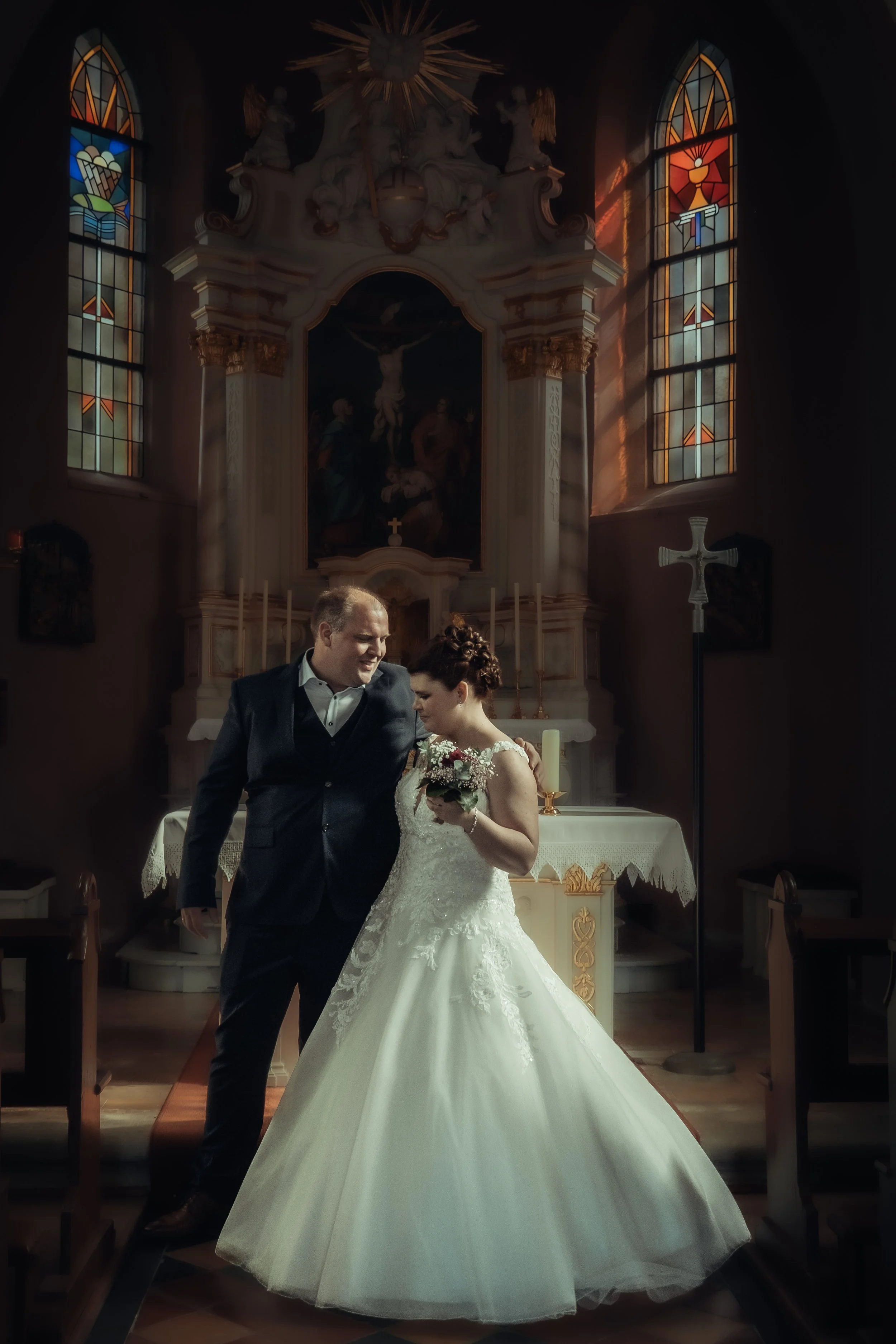 A bride and groom standing together in a church during their wedding ceremony, with stained glass windows and religious symbols in the background.