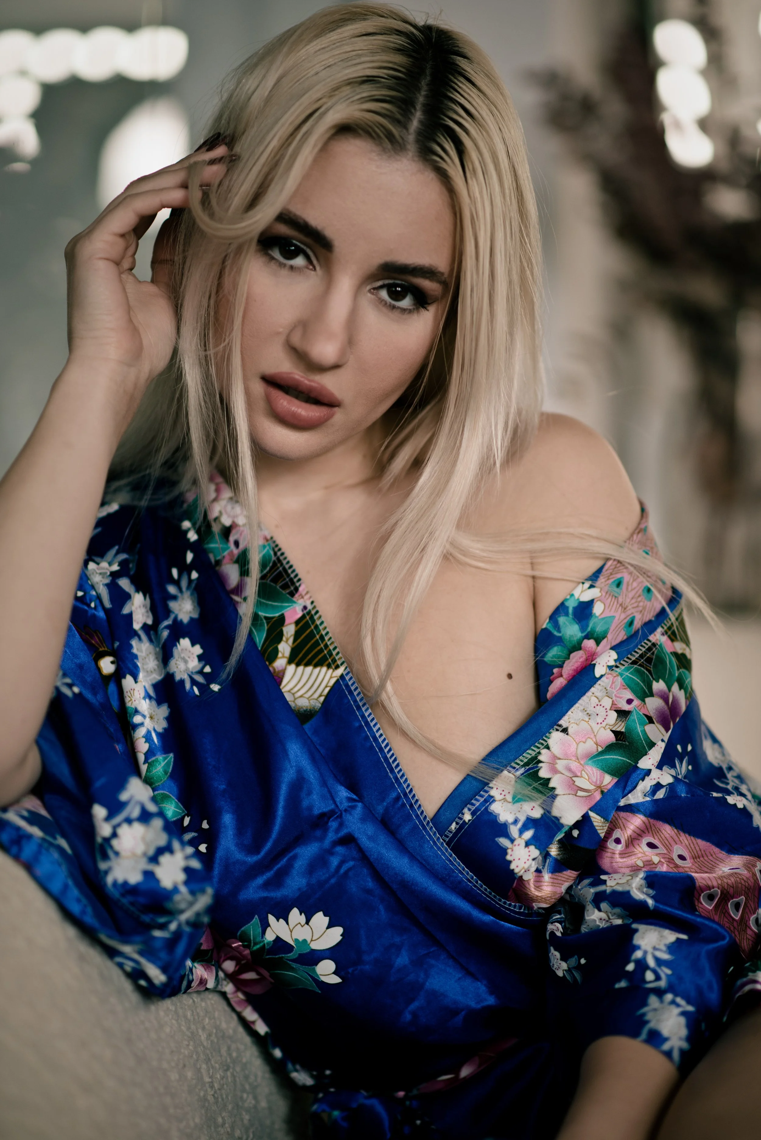 A woman with blonde hair styled in loose waves, wearing makeup, and dressed in a blue floral kimono, sitting on the floor with one hand touching her hair and a slightly open mouth showing a seductive expression.