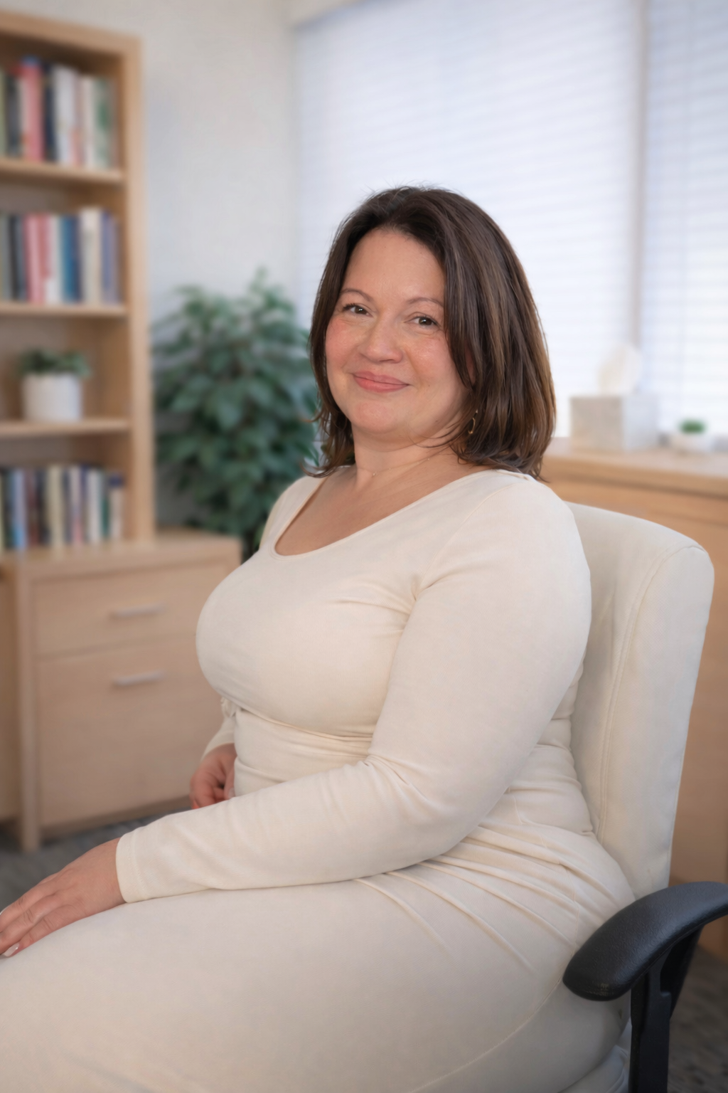 A woman with shoulder-length brown hair sitting in a cream-colored armchair in a well-lit room with a bookshelf and green plant in the background.