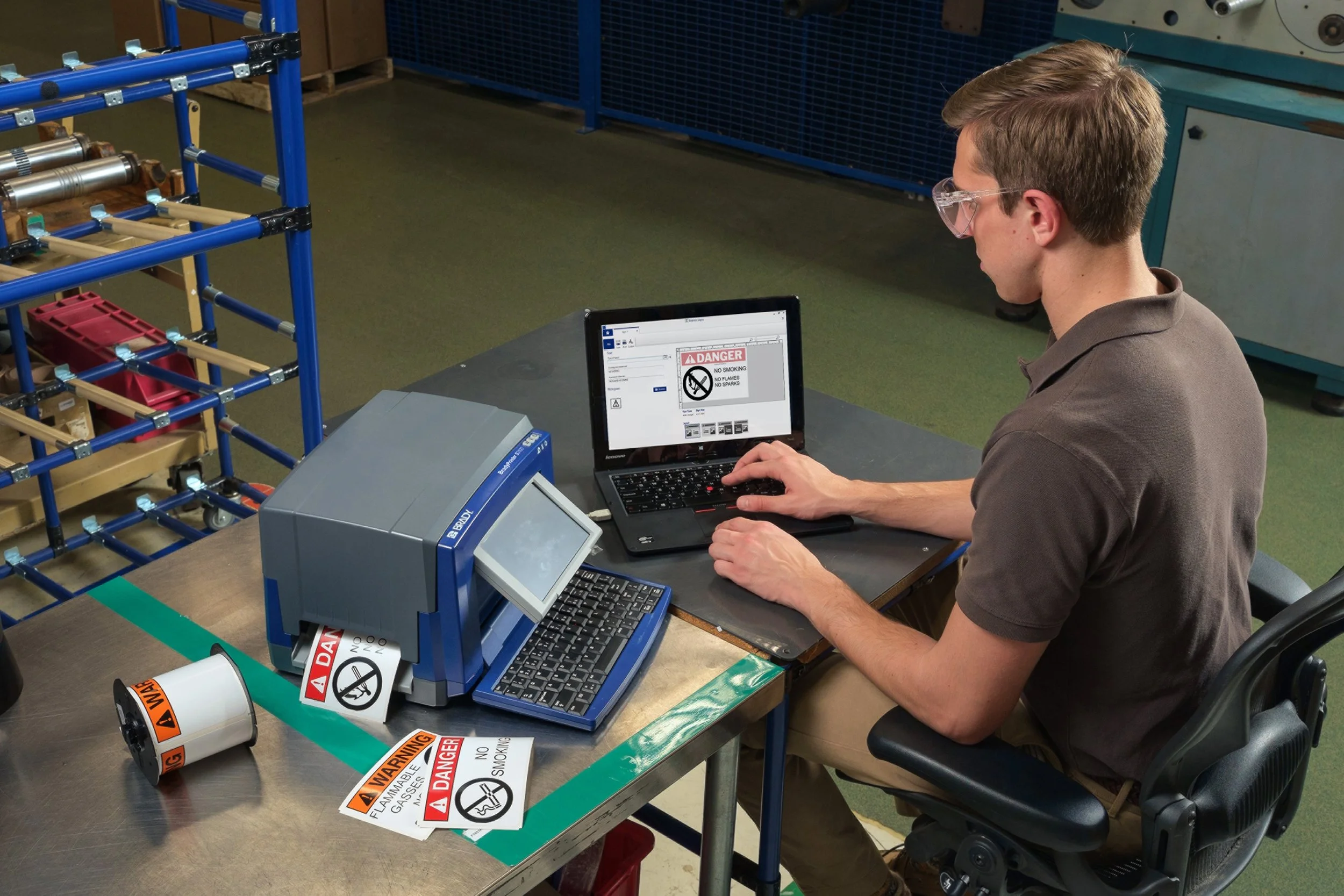 A man wearing safety glasses working with electronic equipment at a workstation that has warning signs and labels, including a label that says "DANGER" and a reel of warning tape with similar signs.