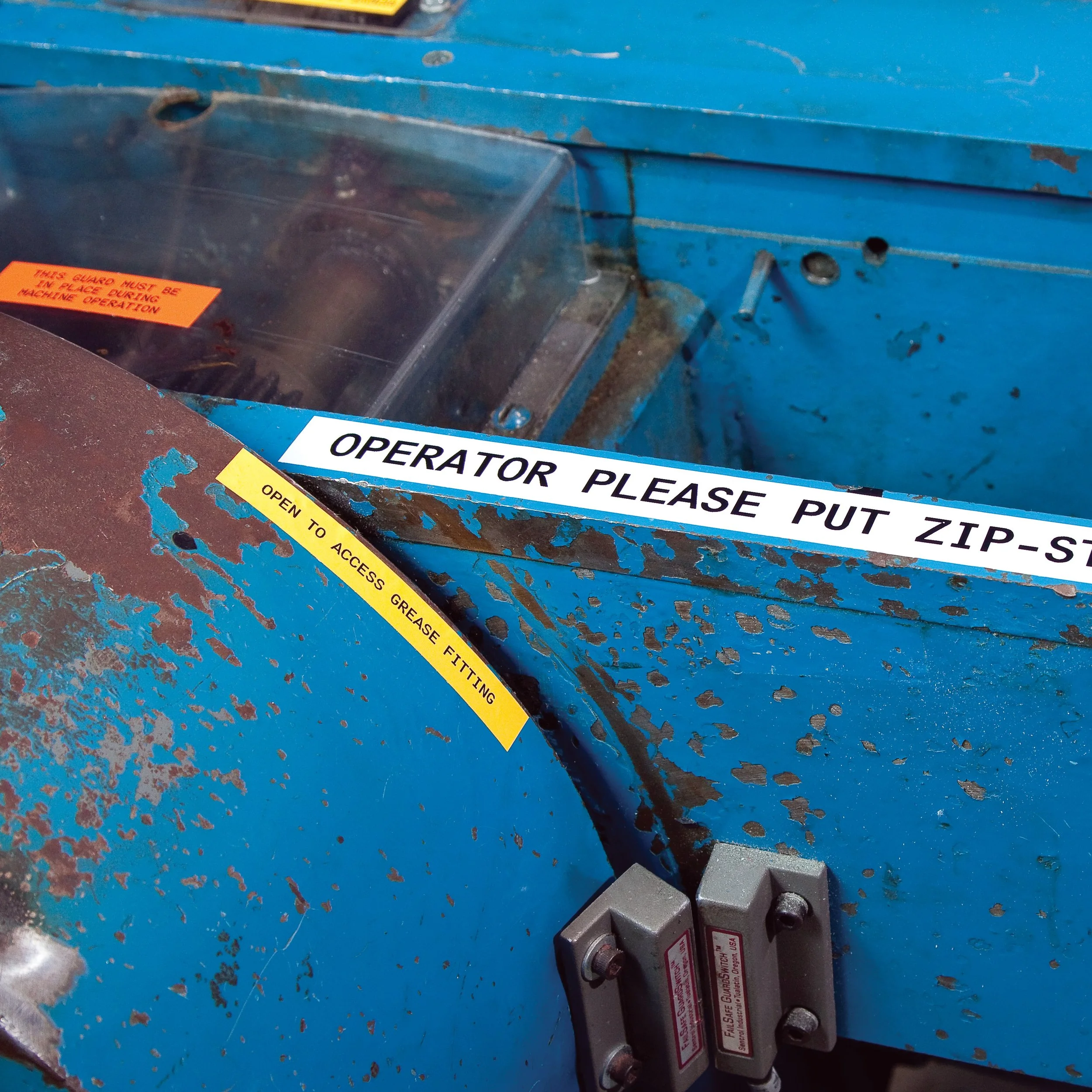 Close-up of worn blue industrial machine with warning and instruction labels, including a sticker that says 'OPERATOR PLEASE PUT ZIP-STOP' and another yellow label that says 'OPEN TO ACCESS GREASE FITTING'.