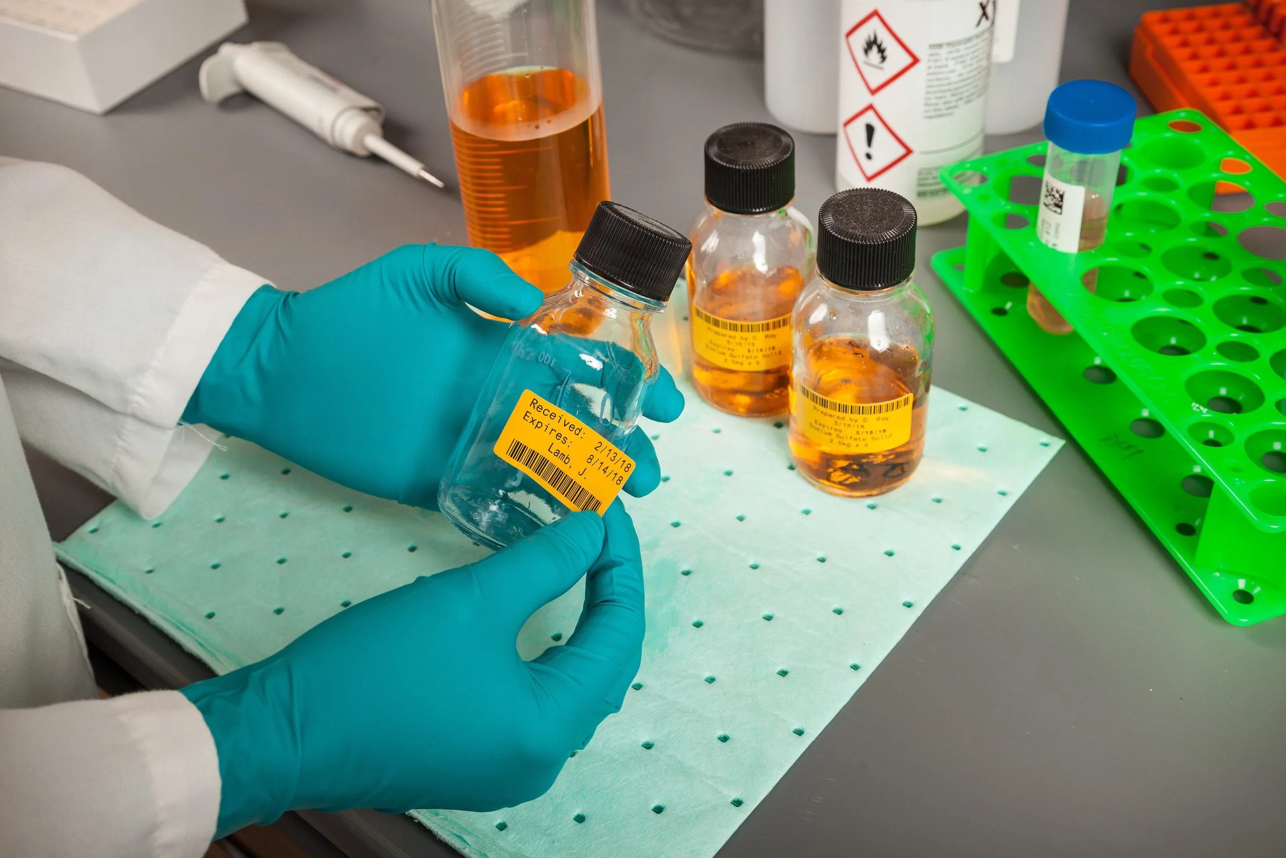 Person wearing blue gloves holding a small bottle with amber liquid in a laboratory setting. The table has more bottles with amber liquid, a beaker, pipette, and a multi-well rack with samples.