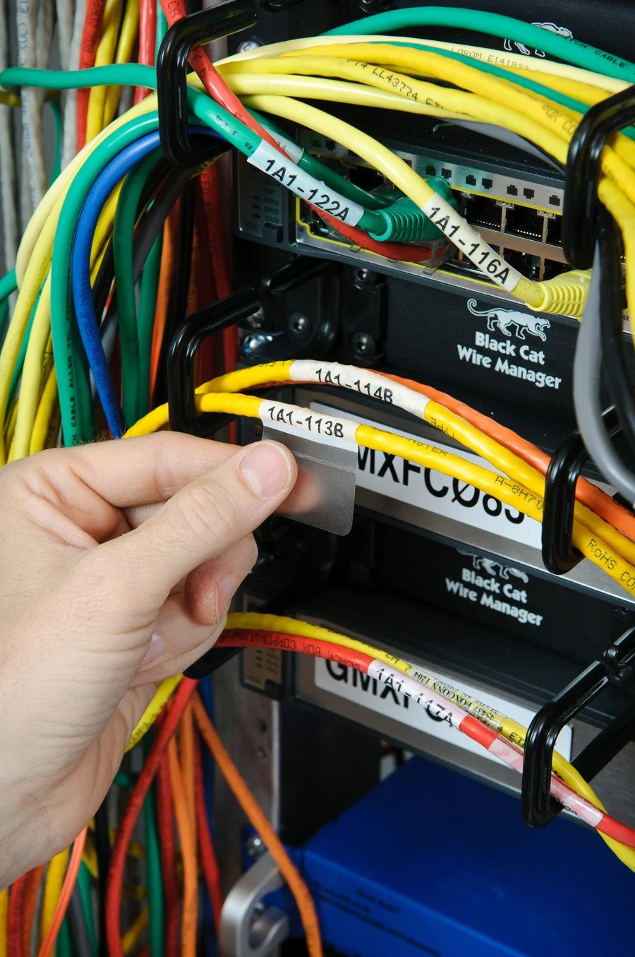 Close-up of a hand holding a cable label in front of a network panel with colored cables and switches. The cables are labeled with white tags containing black text.