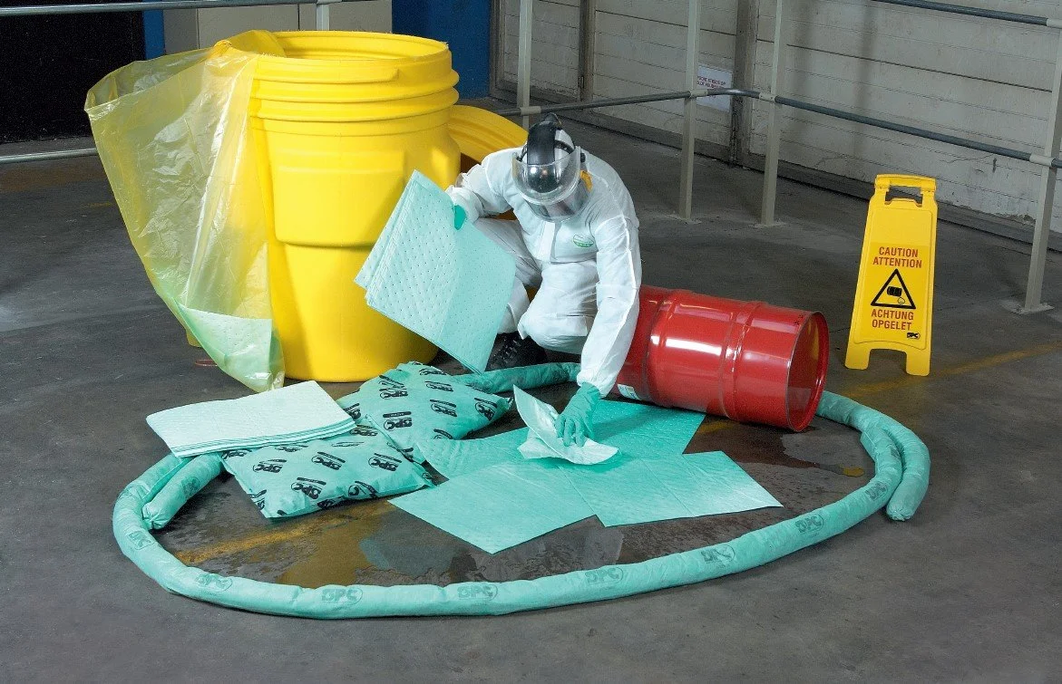 Person dressed in hazmat suit and protective gear cleaning up hazardous spill with absorbent materials in a warehouse.