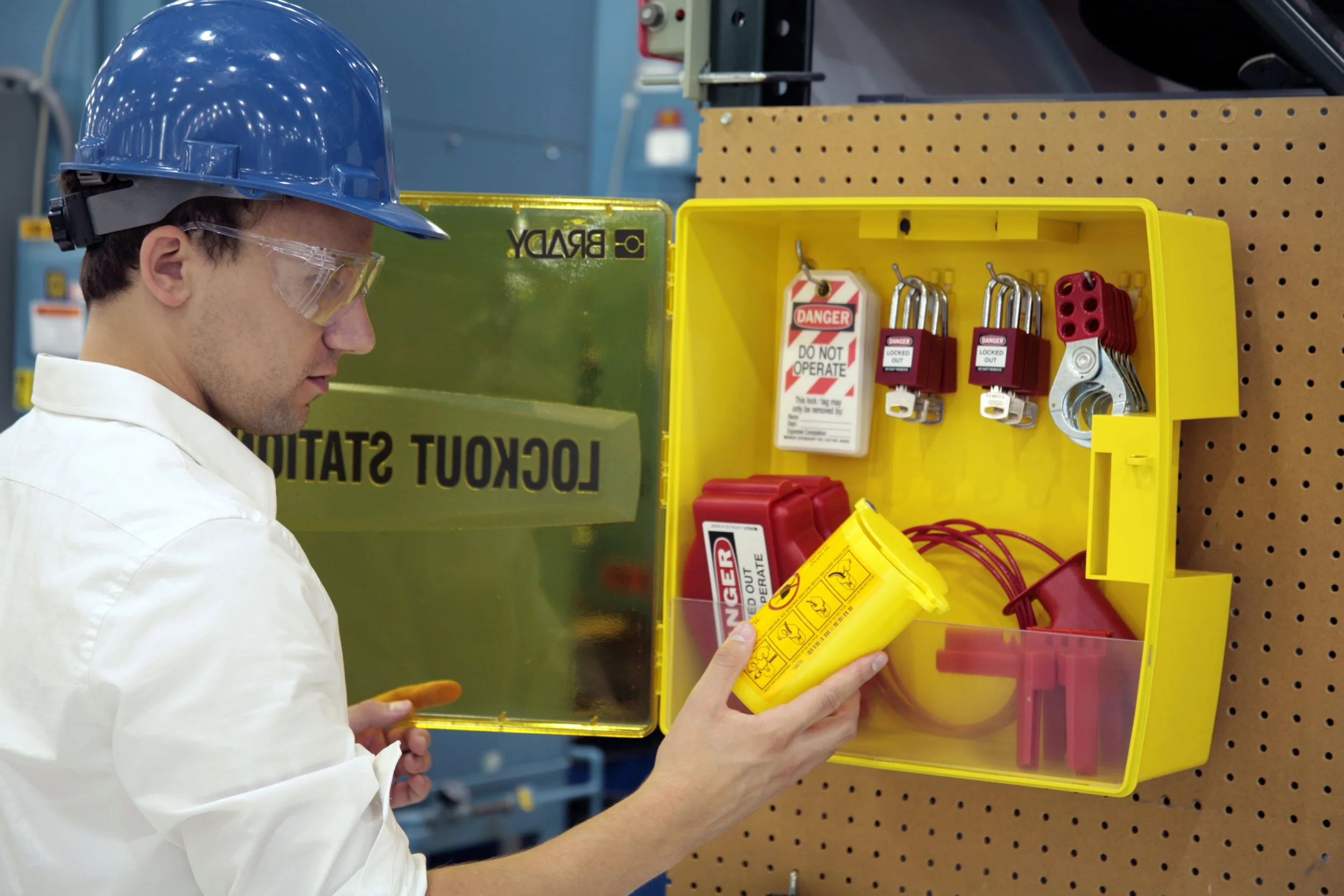 A worker in safety gear, including a blue helmet and safety glasses, inspecting a yellow emergency response kit containing locks, warning tags, and other safety apparatus in a factory setting.