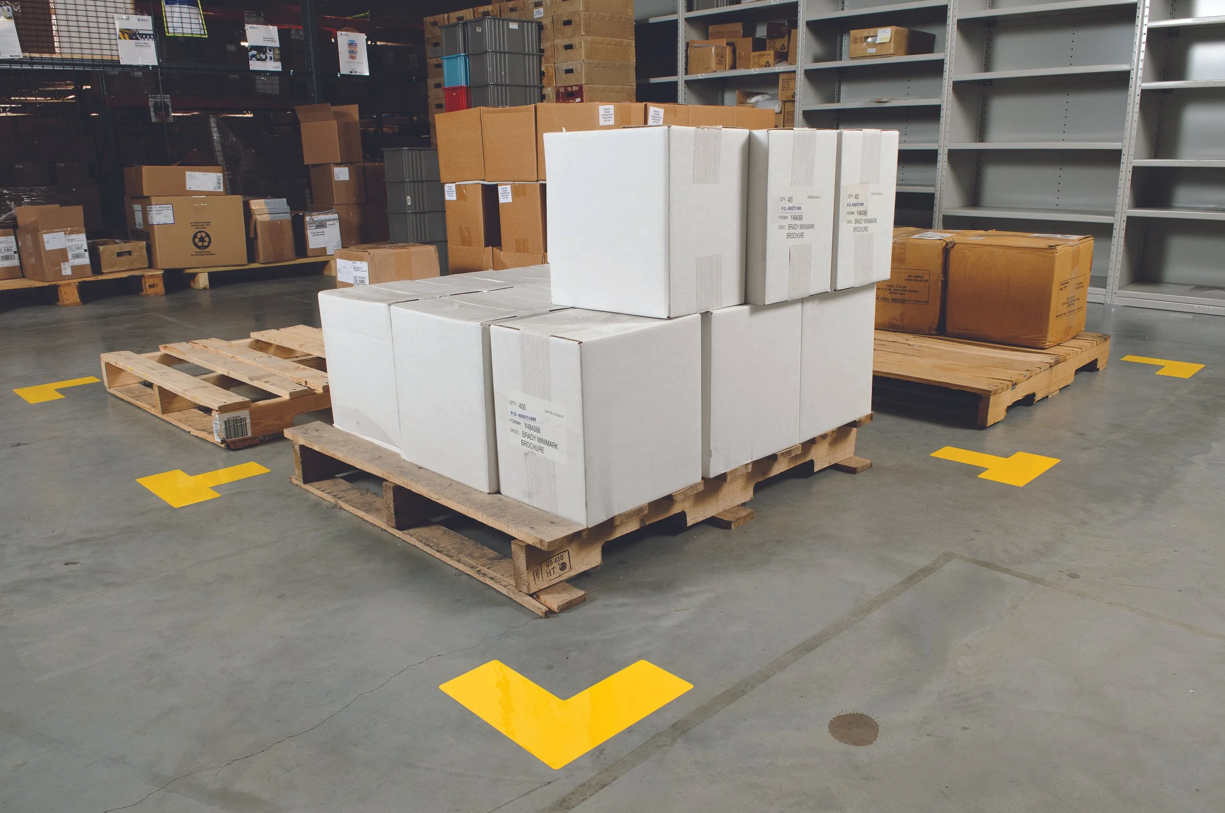 Warehouse with pallets of boxed goods, including white cardboard boxes on a wooden pallet, surrounded by other boxes and shelves, with yellow markings on the concrete floor.