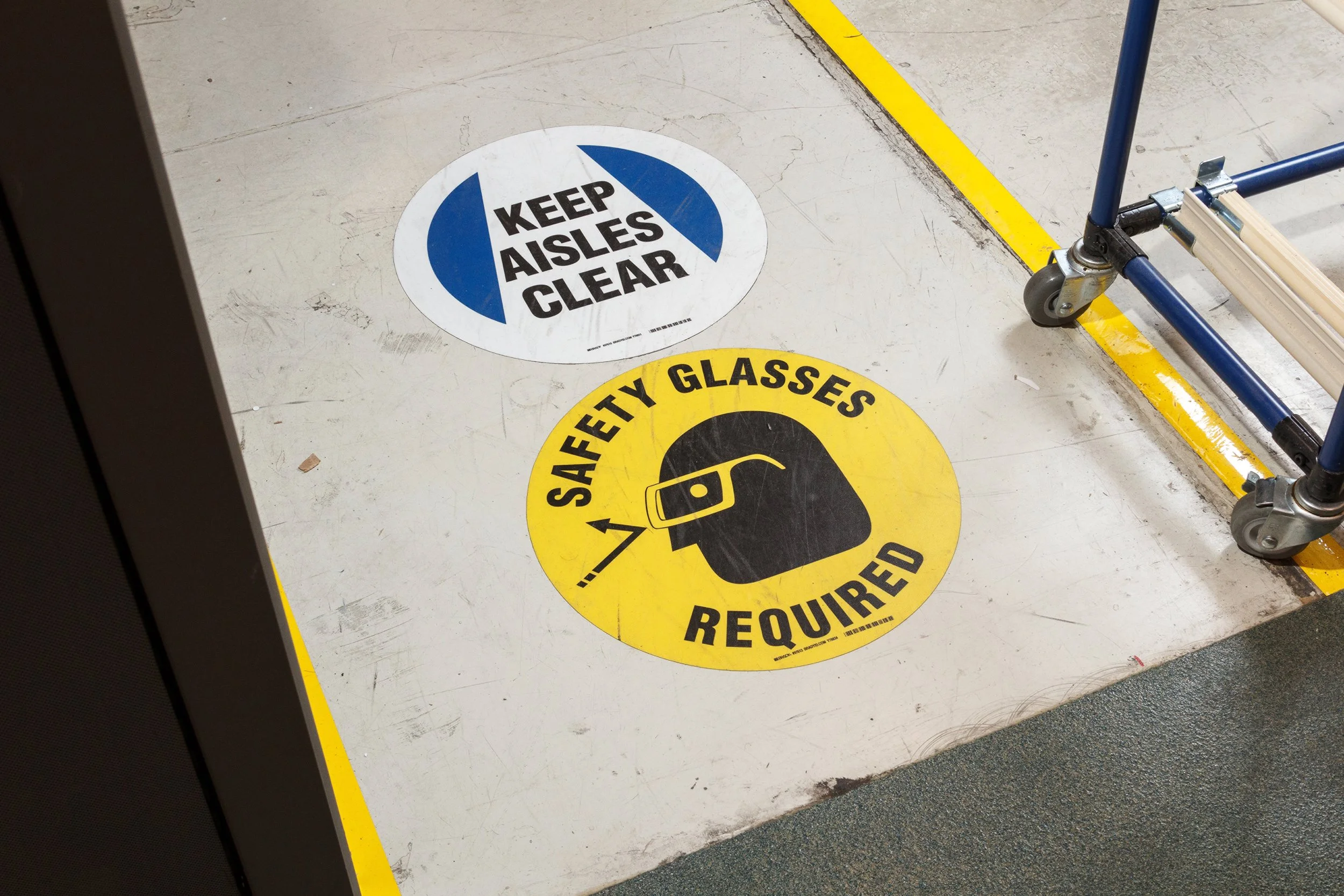 Floor stickers indicating safety requirements; one says 'Keep Aisles Clear' in black text on a white and blue background, the other says 'Safety Glasses Required' with a graphic of safety glasses on a yellow background.