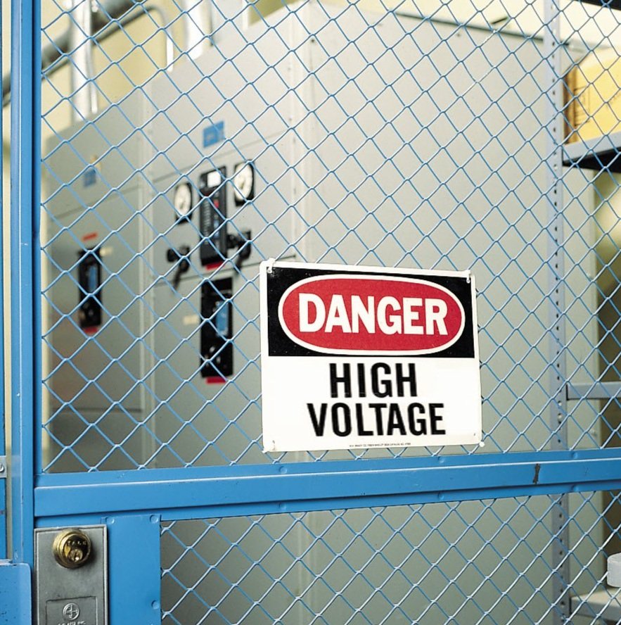 A warning sign on a blue metal fence reads 'DANGER HIGH VOLTAGE', indicating electrical hazard inside. Behind the fence are electrical control panels and equipment.
