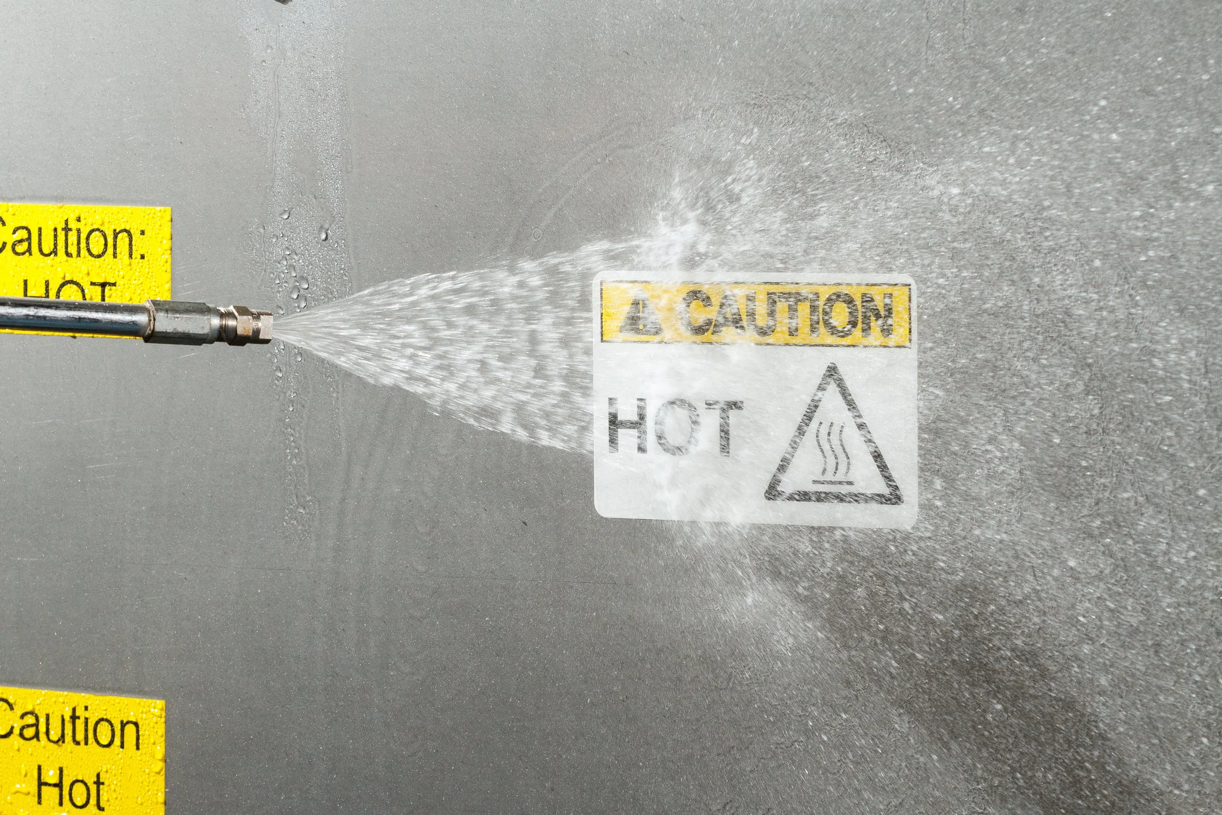 A high-pressure water jet cleaning a surface with caution and hot warning labels.