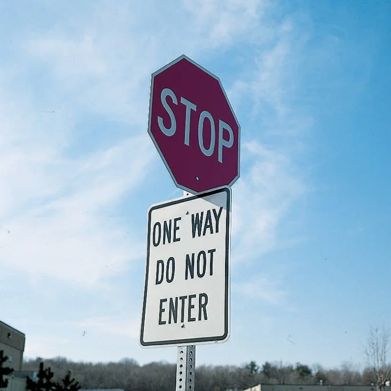 A red stop sign with a white border and a white 'STOP' label, positioned above a white traffic sign that says 'ONE WAY DO NOT ENTER' in black letters, mounted on a metal pole against a blue sky backdrop.