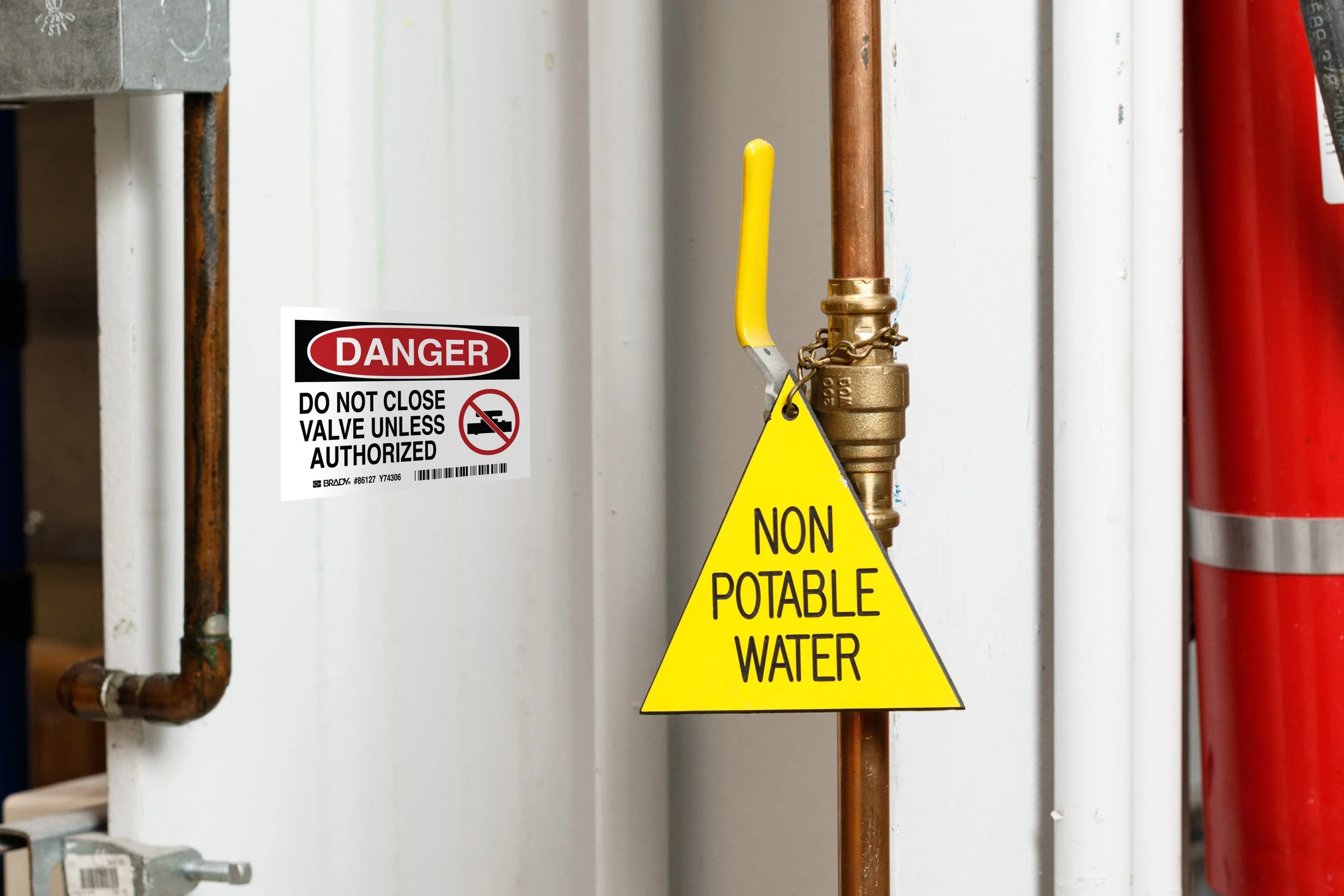 A yellow triangular sign reading 'NON POTABLE WATER' is attached to copper piping. A white sign with a red 'DANGER' label and black text warns not to close the valve unless authorized, indicating the water is non-potable and unsafe to drink.
